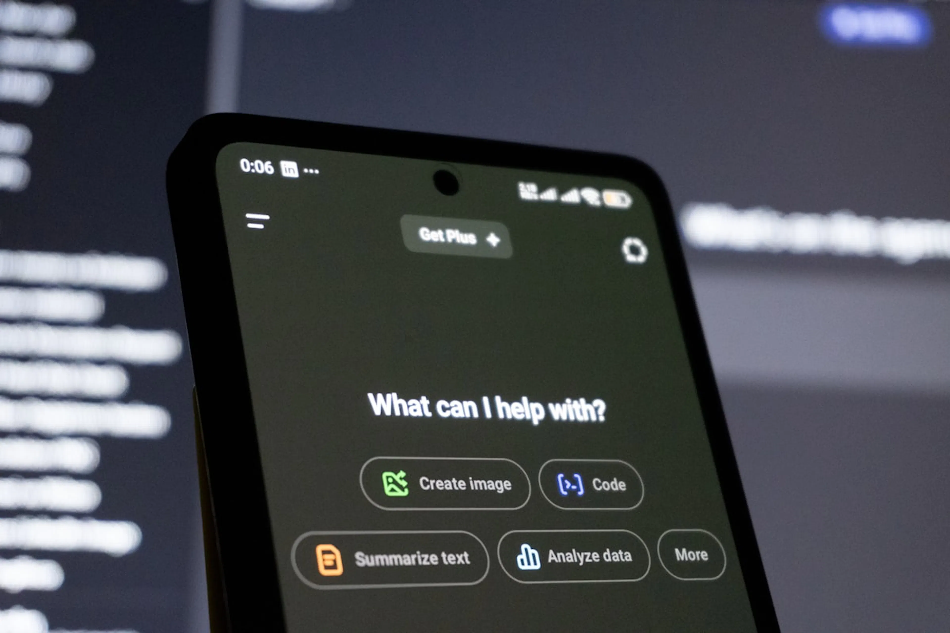 Smartphone held up to a code-filled monitor, showing an AI assistant asking what it can help with