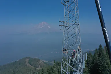 Drone view of a crew working high on a lattice cell tower, with a hazy mountain peak in the distance