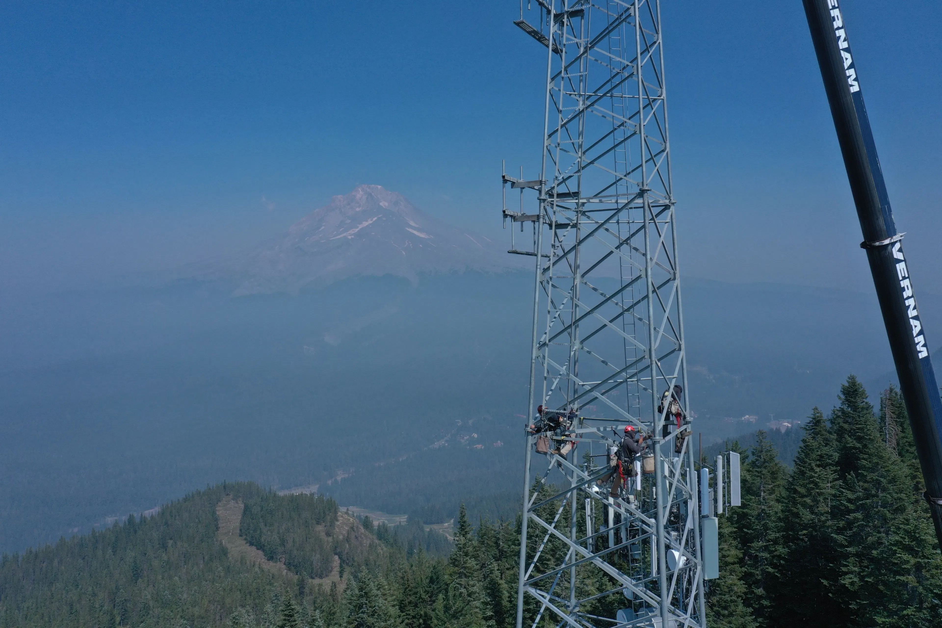 Drone view of a crew working high on a lattice cell tower, with a hazy mountain peak in the distance