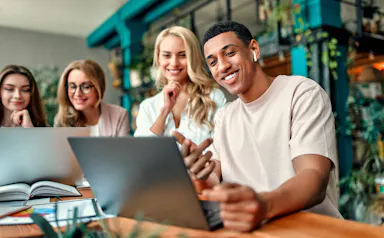 Four coworkers gathered around a laptop in a plant-filled office, smiling at the screen