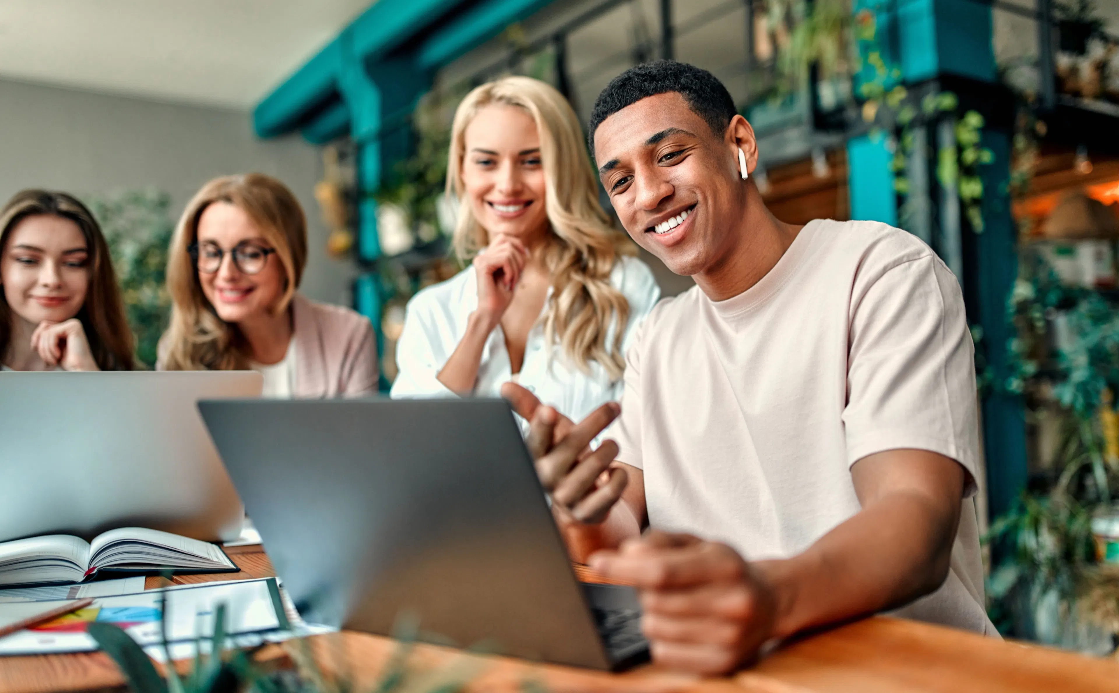 Four coworkers gathered around a laptop in a plant-filled office, smiling at the screen