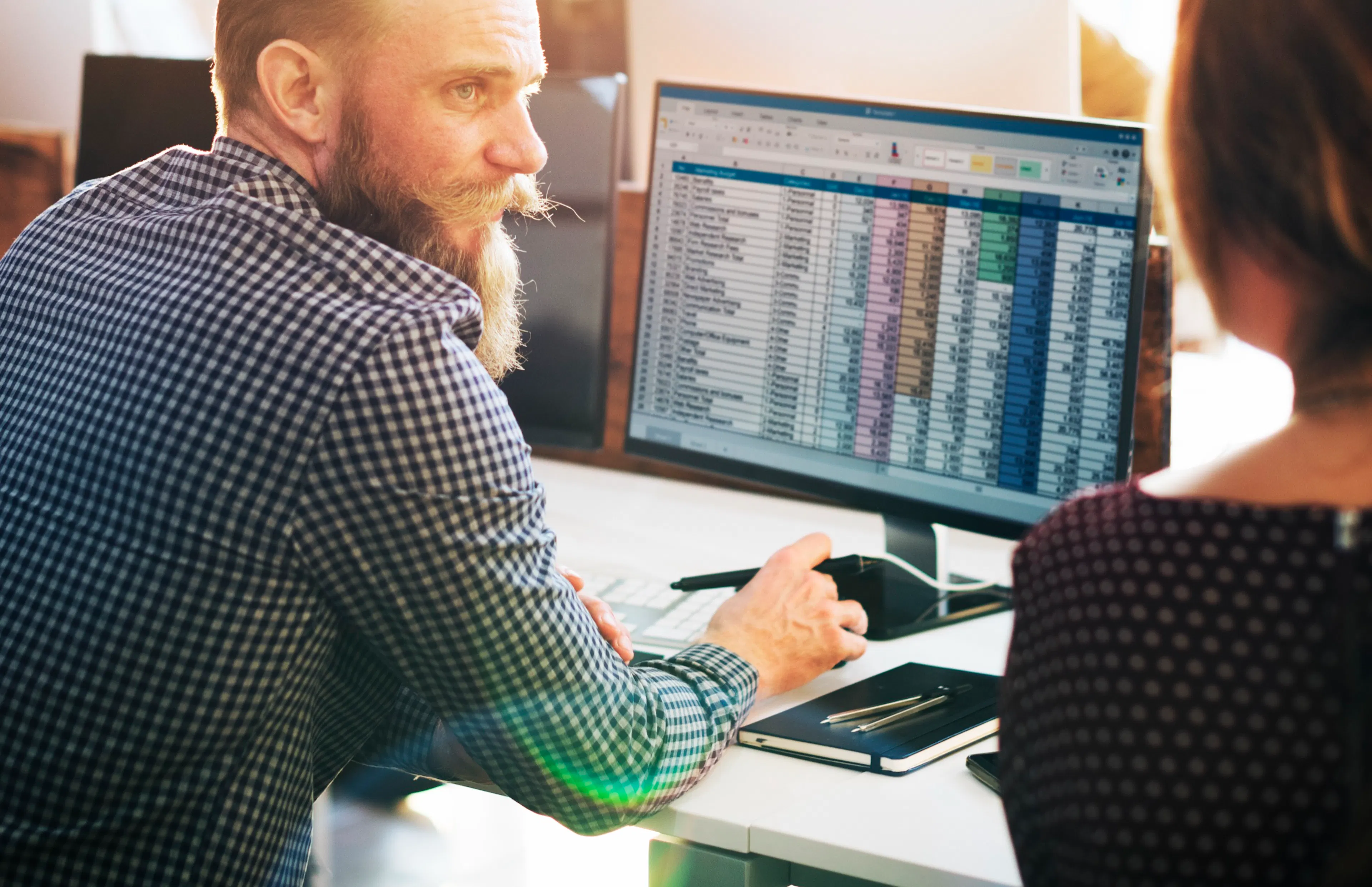 Two colleagues at a desk reviewing a dense color-coded spreadsheet on a monitor