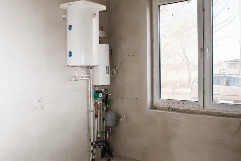 Water heater with pipes and valves mounted on a white wall near a window.