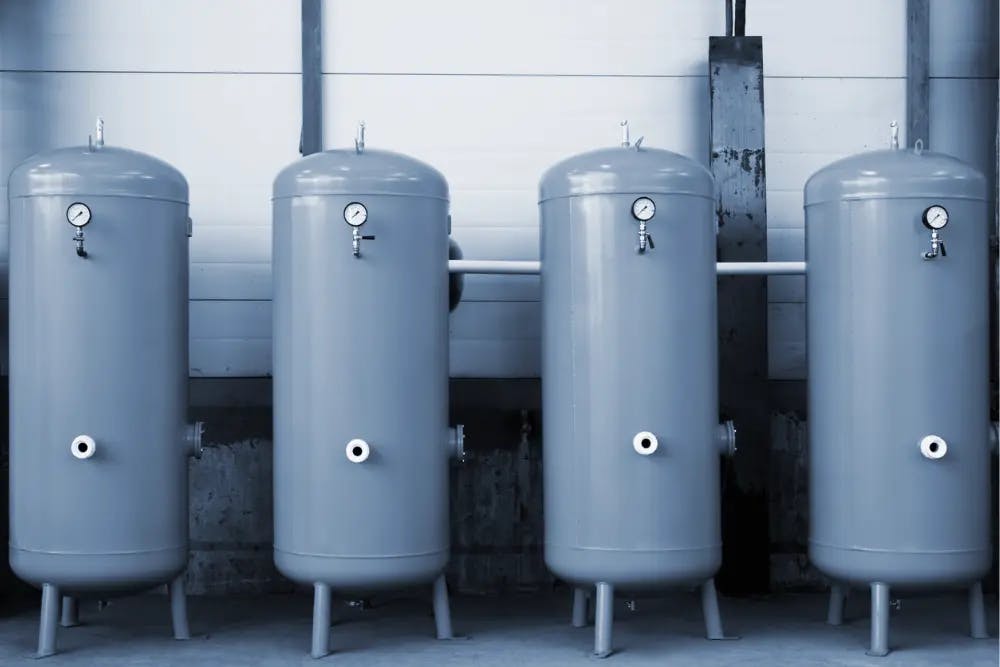 Row of four grey hot water tanks with pressure gauges mounted on a wall
