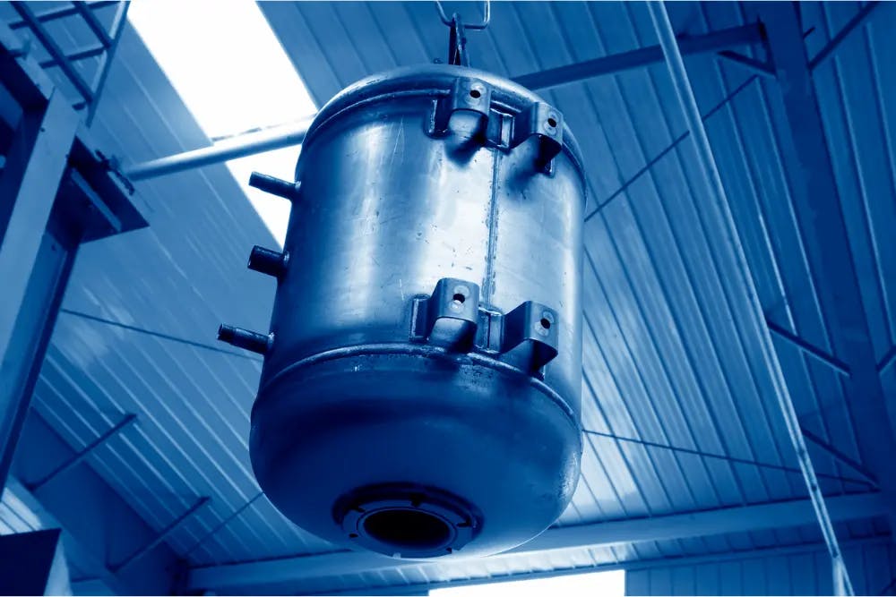 A large hot water tank mounted on the ceiling, with mounting brackets and ports, photographed from below with a blue tint.