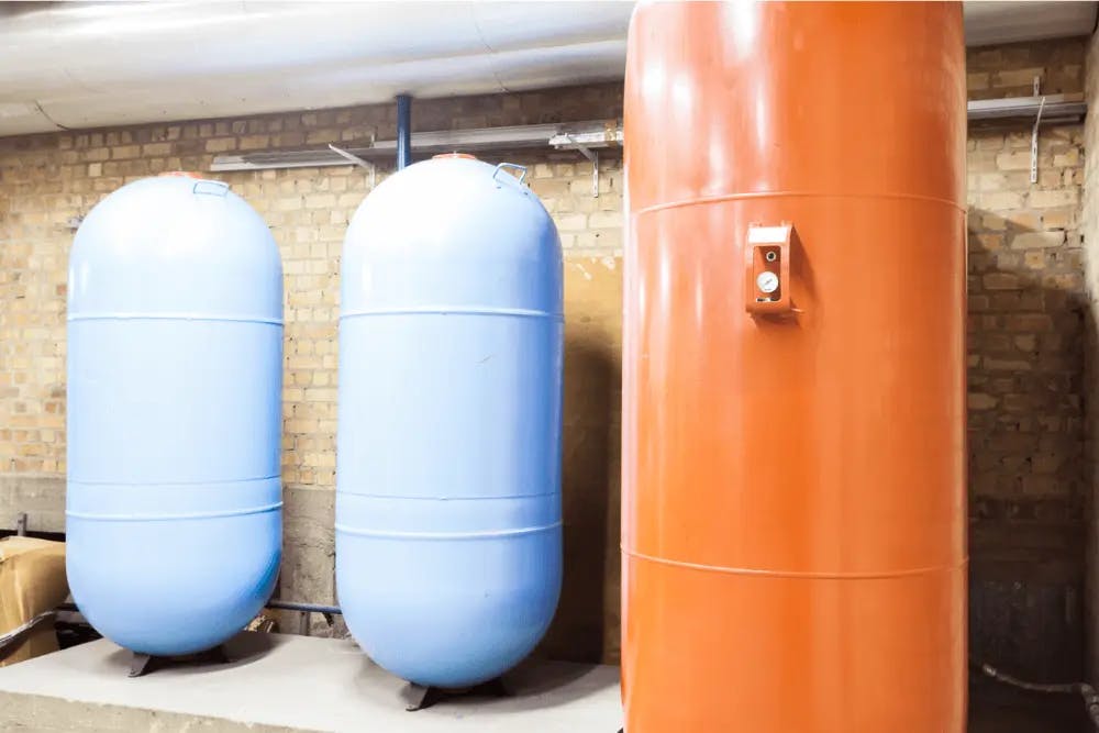 Two blue hot water storage tanks and one larger orange tank with control panel, mounted against brick wall in mechanical room