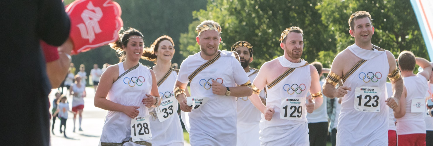 A group of people dressed in togas wearing Mace Foundation running bibs with the Olympic rings above them