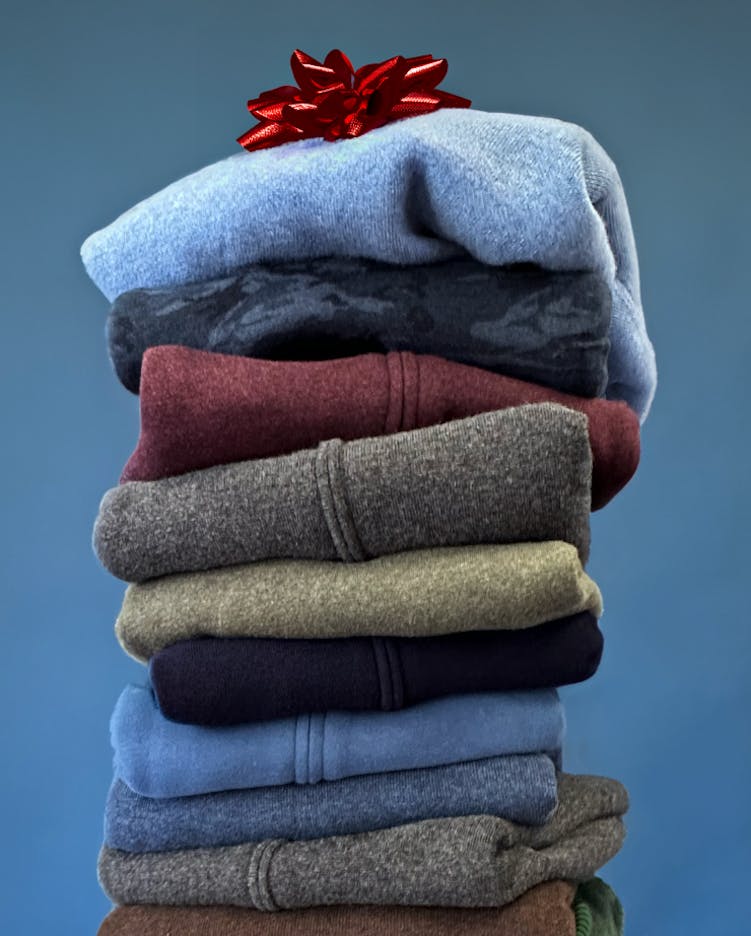 Folded and stacked mack Weldon sweaters with a red holiday bow on top and a medium blue background