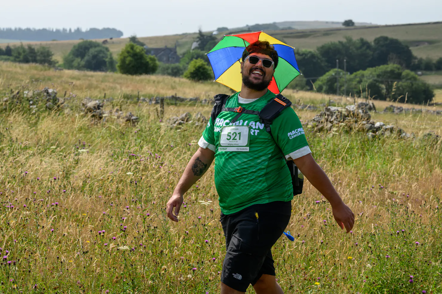 One male hiker walking through grass, wearing a green Macmillan top and a rainbow umbrella hat.