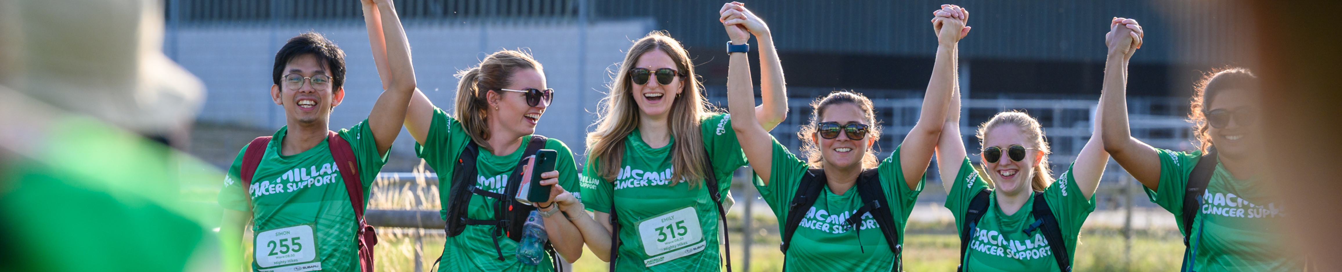 A group of 6 hikers holding hands and cheering, all wearing Macmillan tops.