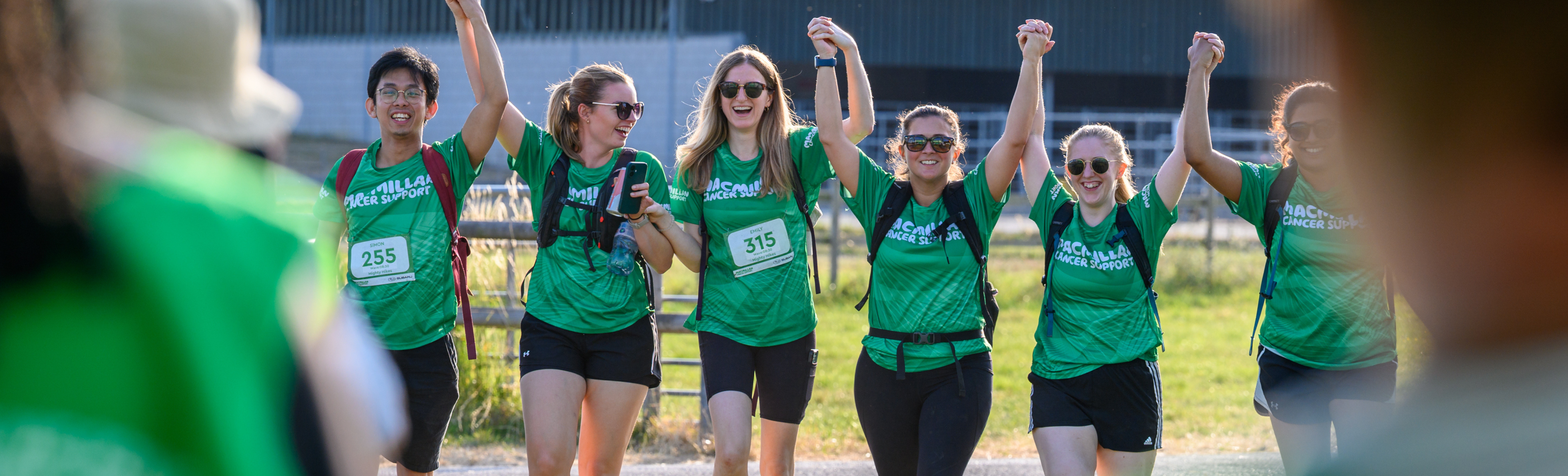 A group of 6 hikers holding hands and cheering, all wearing Macmillan tops.