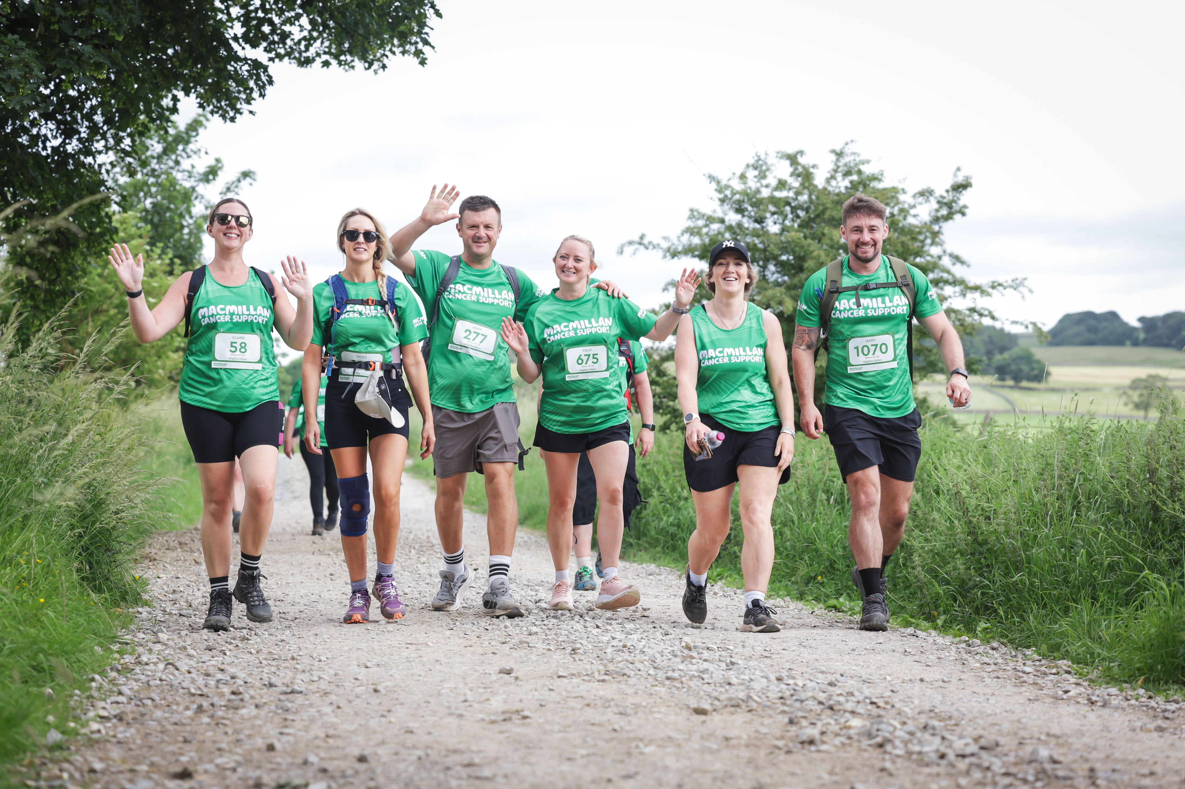 A group of hikers wearing Macmillan tops, smiling and waving at the camera.
