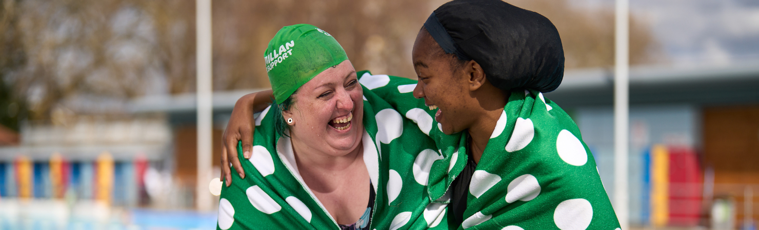 Two women are stood laughing together in a lido setting after just completing a swim.    The woman on the left is wearing a green Macmillan swimming cap and has a green and white polka dot towel wrapped around her shoulders.    The woman on the right has her arm around the other with her hand resting on her shoulder. She is wearing a black soul cap and has a green and Macmillan towel wrapped around her shoulders as well.
