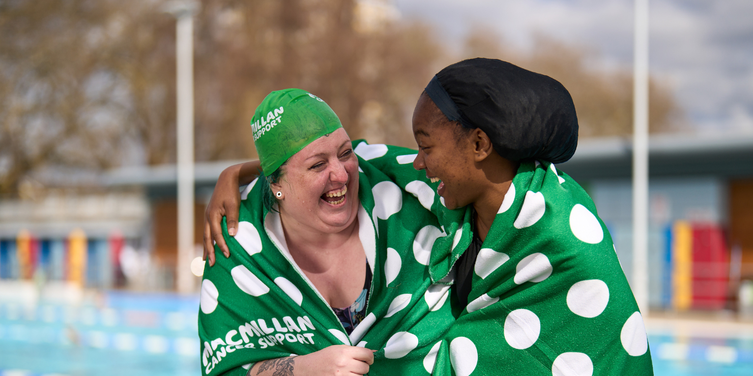Two women are stood laughing together in a lido setting after just completing a swim.    The woman on the left is wearing a green Macmillan swimming cap and has a green and white polka dot towel wrapped around her shoulders.    The woman on the right has her arm around the other with her hand resting on her shoulder. She is wearing a black soul cap and has a green and Macmillan towel wrapped around her shoulders as well.