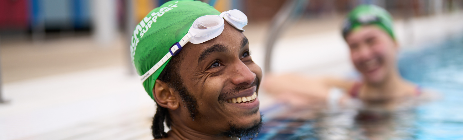 A close up of a young man in the water resting against the swimming pool edge.    He wears a green Macmillan swimming cap and white googles (resting on his forehead). He is looking up and smiling off centre, as if talking to someone to the right of the camera. The rest of the image is blurred out, with a young women also resting against the pool edge smiling, wearing a green Macmillan swimming cap and black googles. 