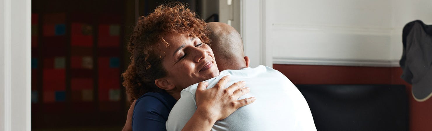 A close up of a female nurse and patient hugging in a waiting room setting. The nurse has auburn curly hair and is facing the camera, eyes closed and a warm smile on her face. The patient is mostly hidden from view but is wearing a white T-shirt and has lost their hair.
