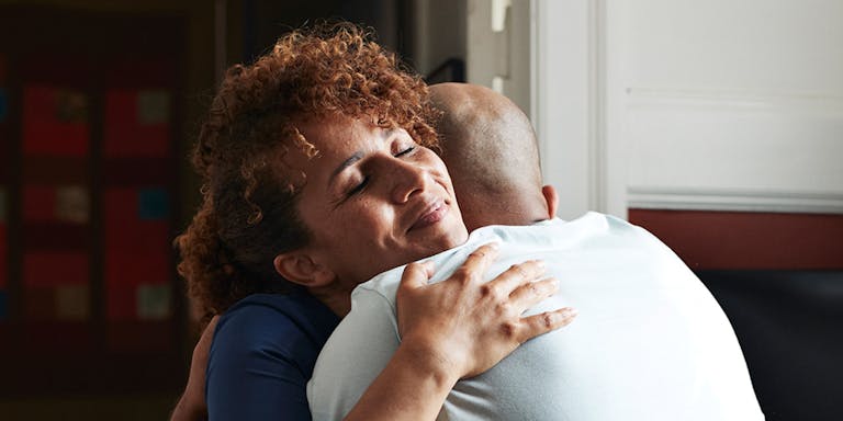 A close up of a female nurse and patient hugging in a waiting room setting. The nurse has auburn curly hair and is facing the camera, eyes closed and a warm smile on her face. The patient is mostly hidden from view but is wearing a white T-shirt and has lost their hair.
