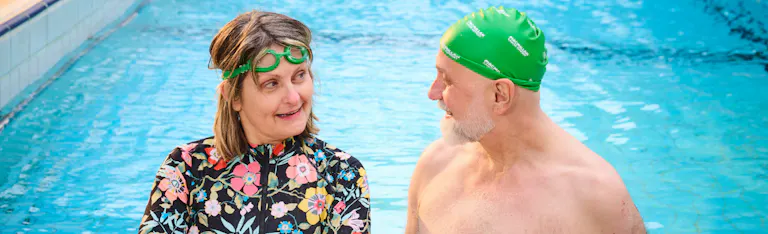 Two swimmers smiling at one another in the pool, one wearing a Macmillan swim cap.