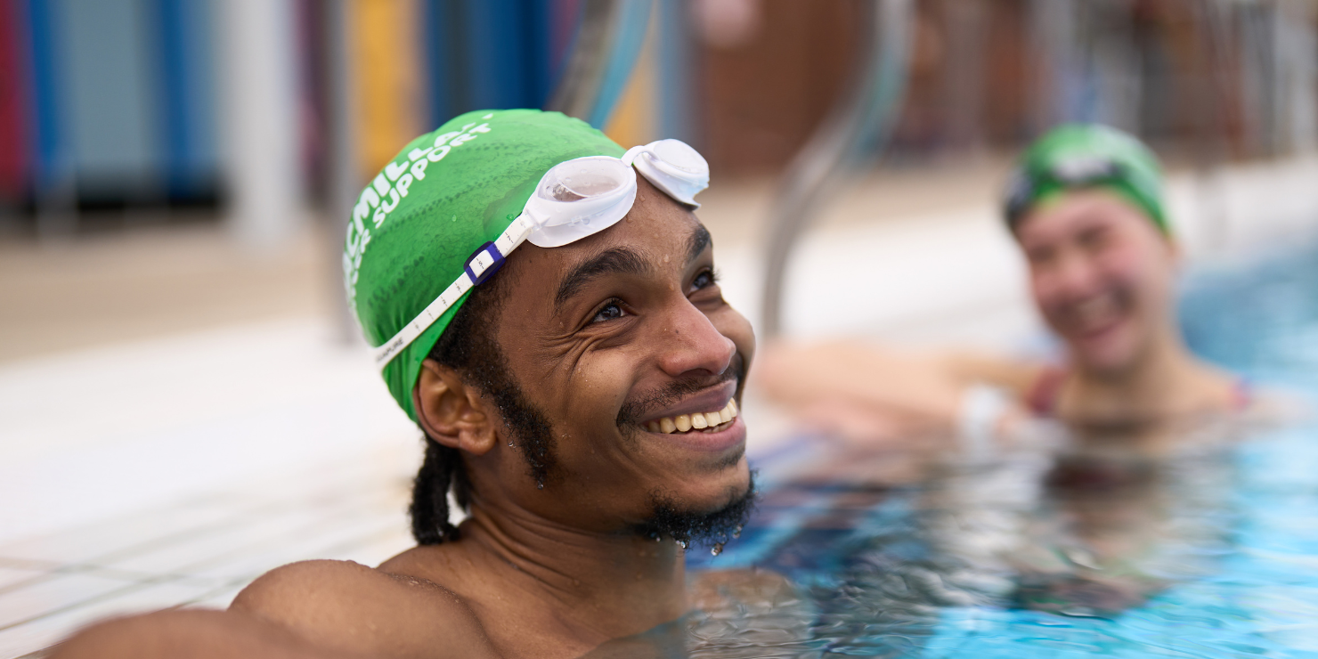 A close up of a young man in the water resting against the swimming pool edge.    He wears a green Macmillan swimming cap and white googles (resting on his forehead). He is looking up and smiling off centre, as if talking to someone to the right of the camera. The rest of the image is blurred out, with a young women also resting against the pool edge smiling, wearing a green Macmillan swimming cap and black googles. 