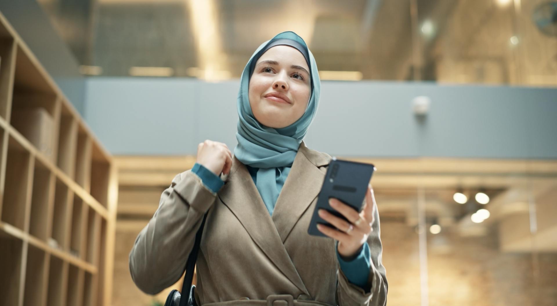 A young woman wearing a hijab walking through a modern office holding her phone