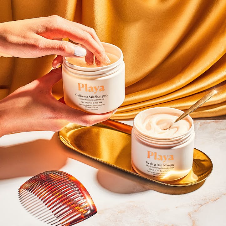 Image of woman's hand dipping into jar of Playa California Salt Shampoo sitting on top a gold plated tray.