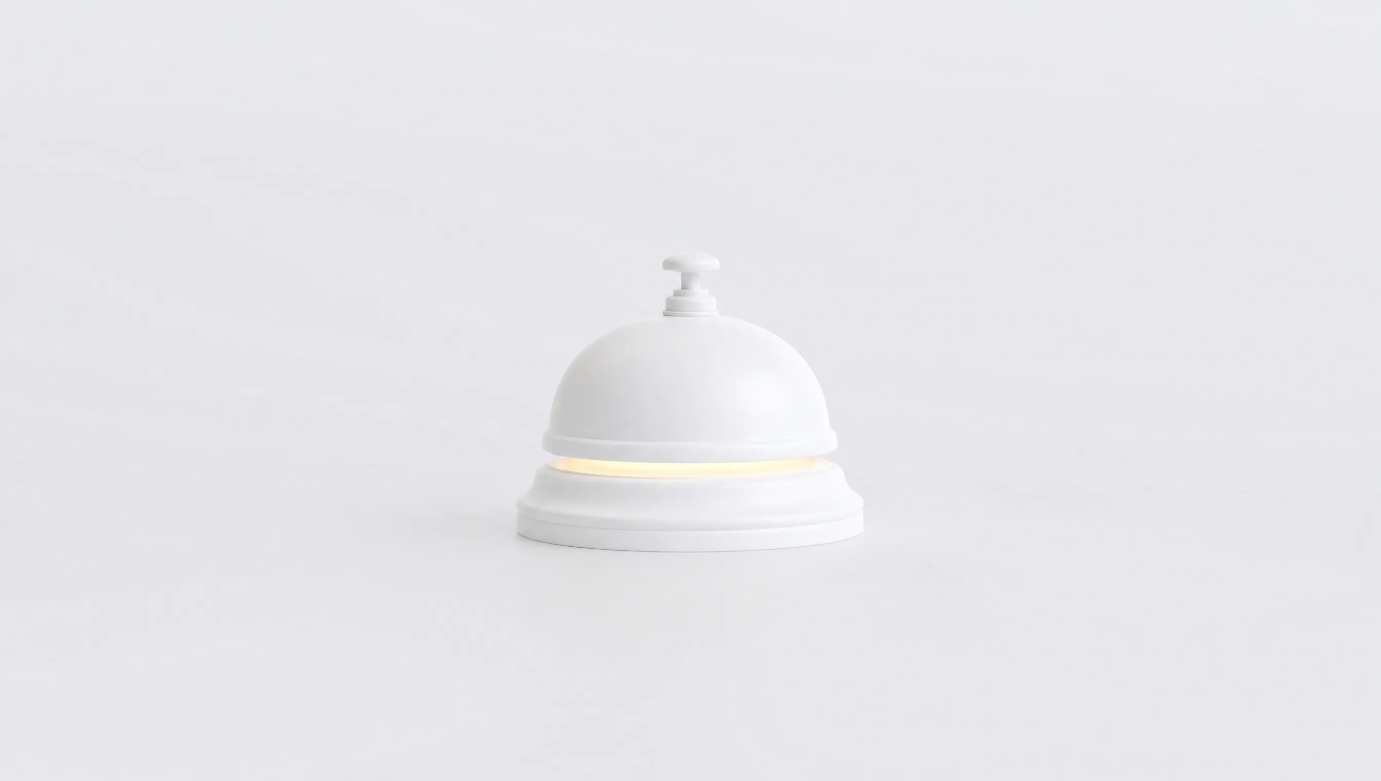 Un campanello da reception scolpito in materiale bianco opaco, visto frontale al centro di un fondo chiaro e vuoto, con un tenue bagliore ambra che filtra dalla fessura fra la campana e la base