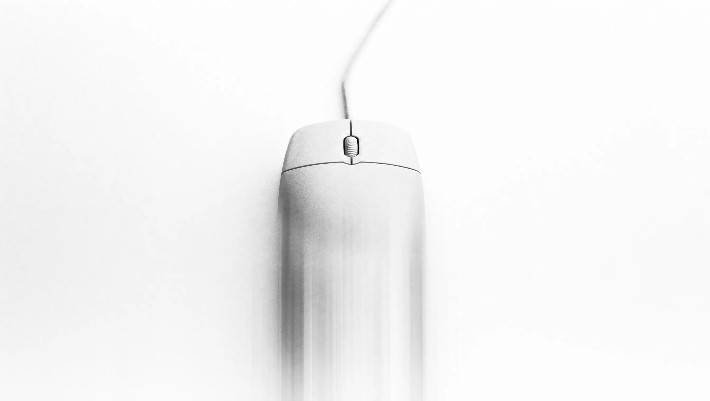 Fotografia in bianco e nero a lunga esposizione di un mouse bianco con filo, visto dall'alto al centro su fondo quasi bianco: i due tasti e la rotella restano nitidi, mentre la parte posteriore si dissolve in scie verticali verso il basso