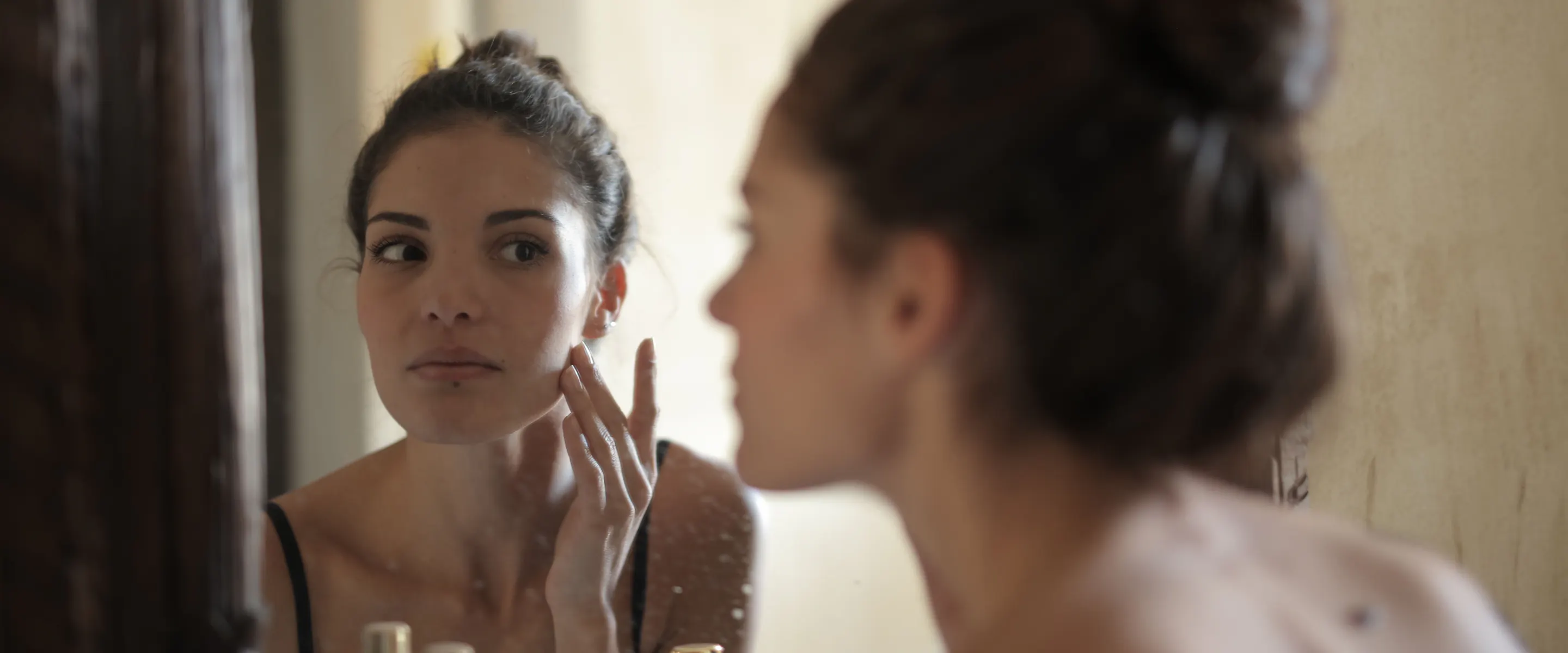 Une jeune adulte observe sa peau dans un miroir, en lumière naturelle.