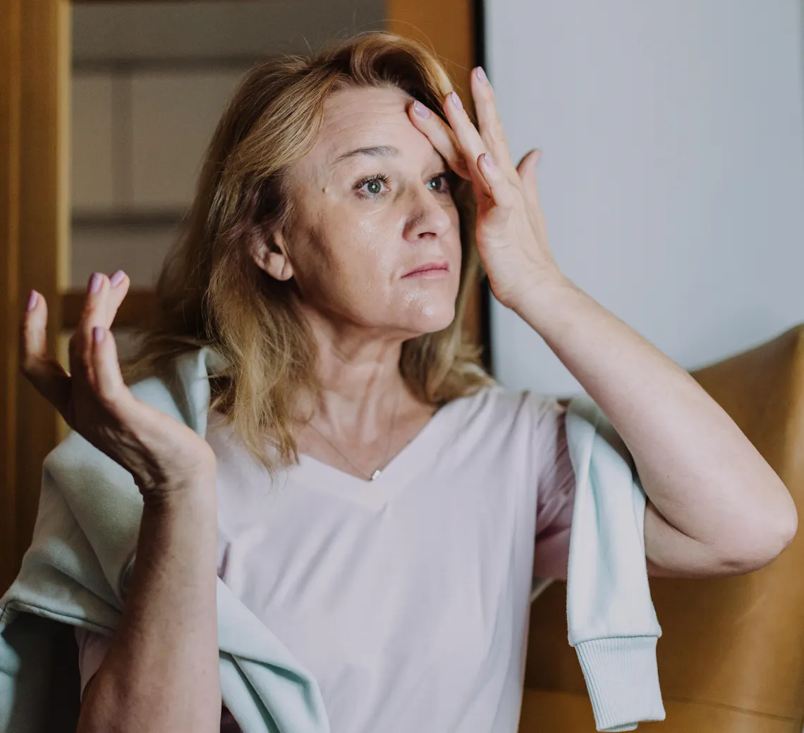 Une femme mature observe son visage, assise dans un fauteuil chez elle.