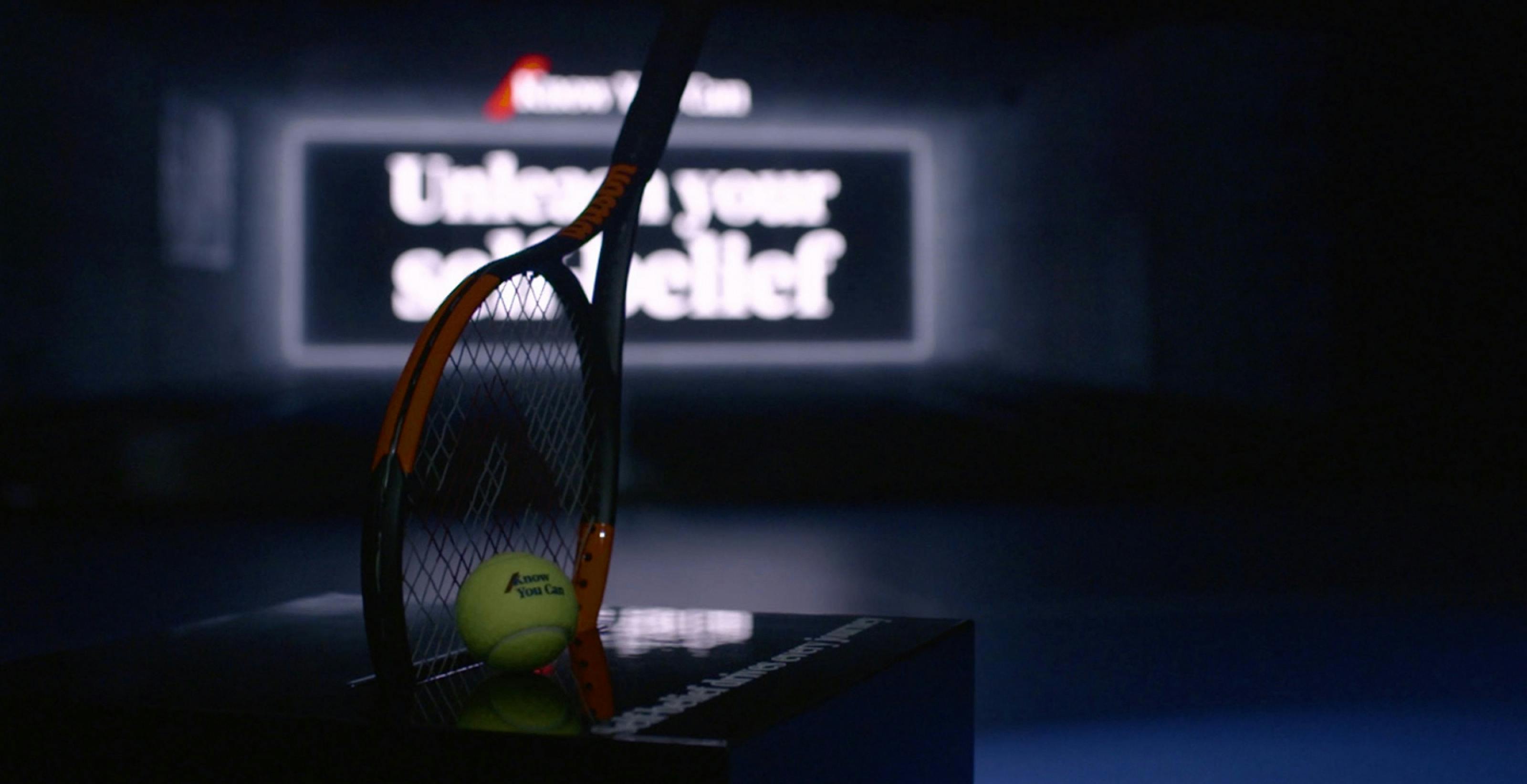 In the foreground, there are a tennis racket and a ball. In the background, the theme of the installation is showed on the screen.