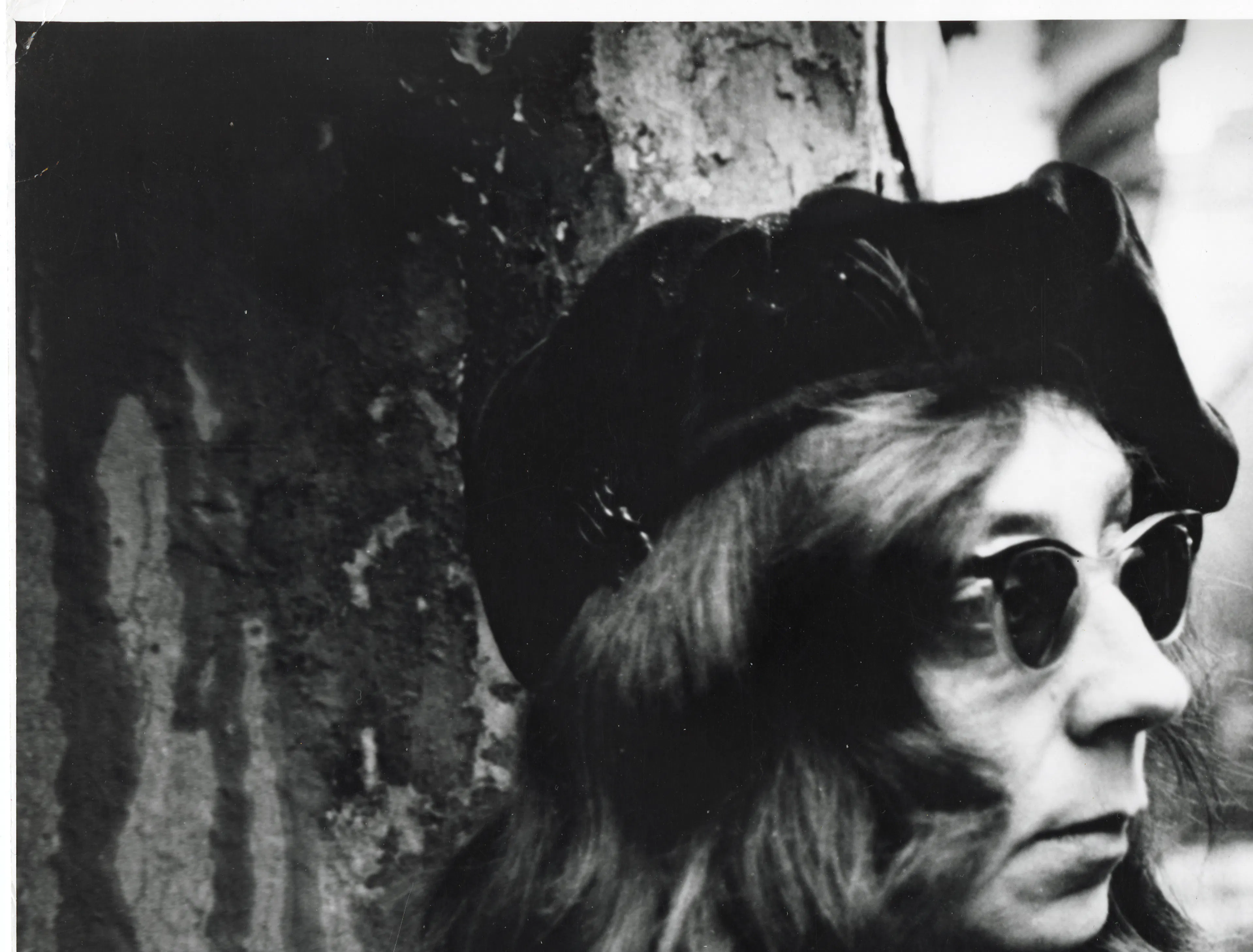 The photograph shows a woman wearing a beret and sunglasses standing behind a tree.