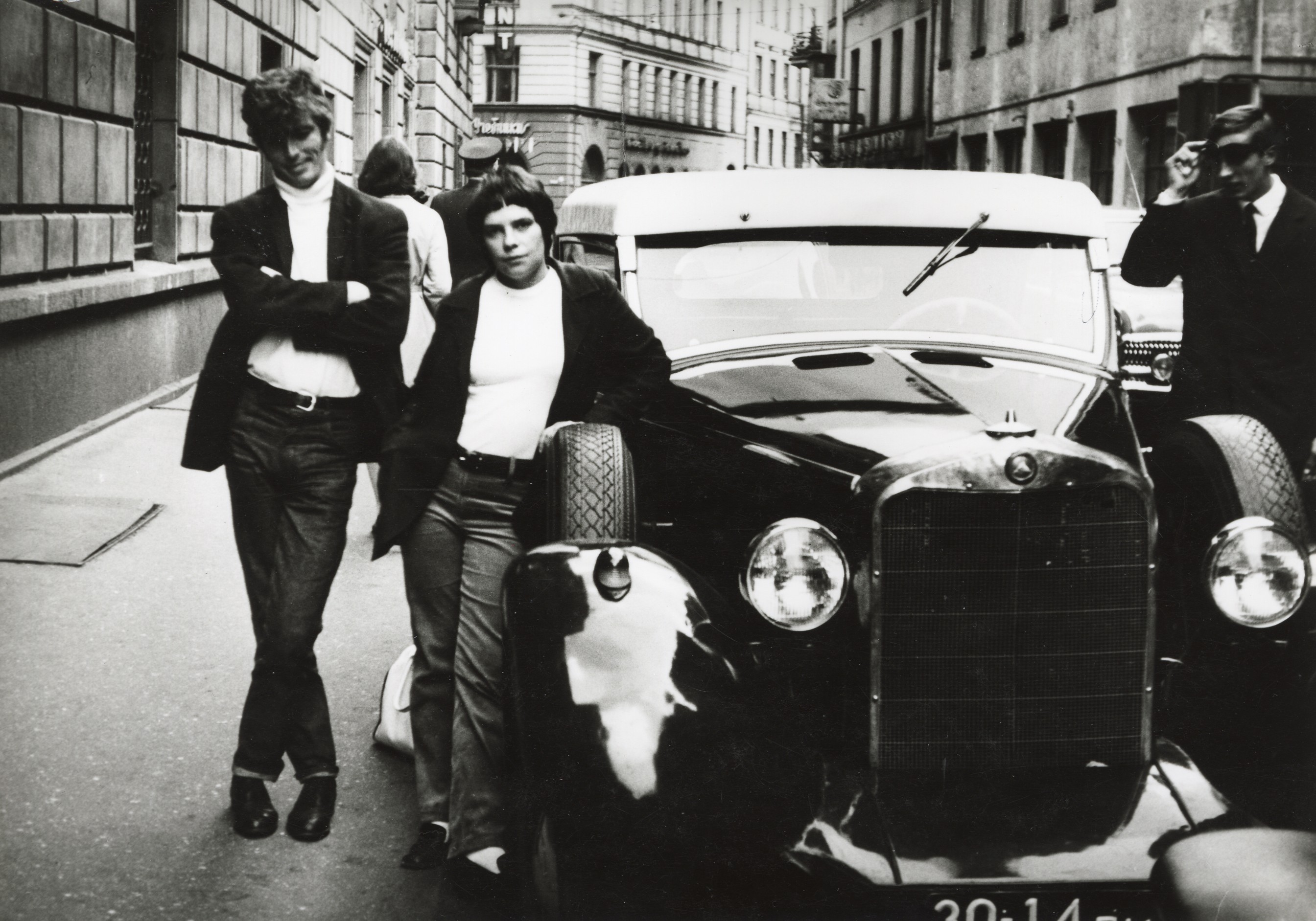 Fotogrāfijā redzams vīrietis un sieviete līdzīgos džemperos un žaketēs. Sieviete ir atspiedusies pret retro automašīnas riepu. Automašīnas kreisajā pusē stāv vīrietis uzvalkā.