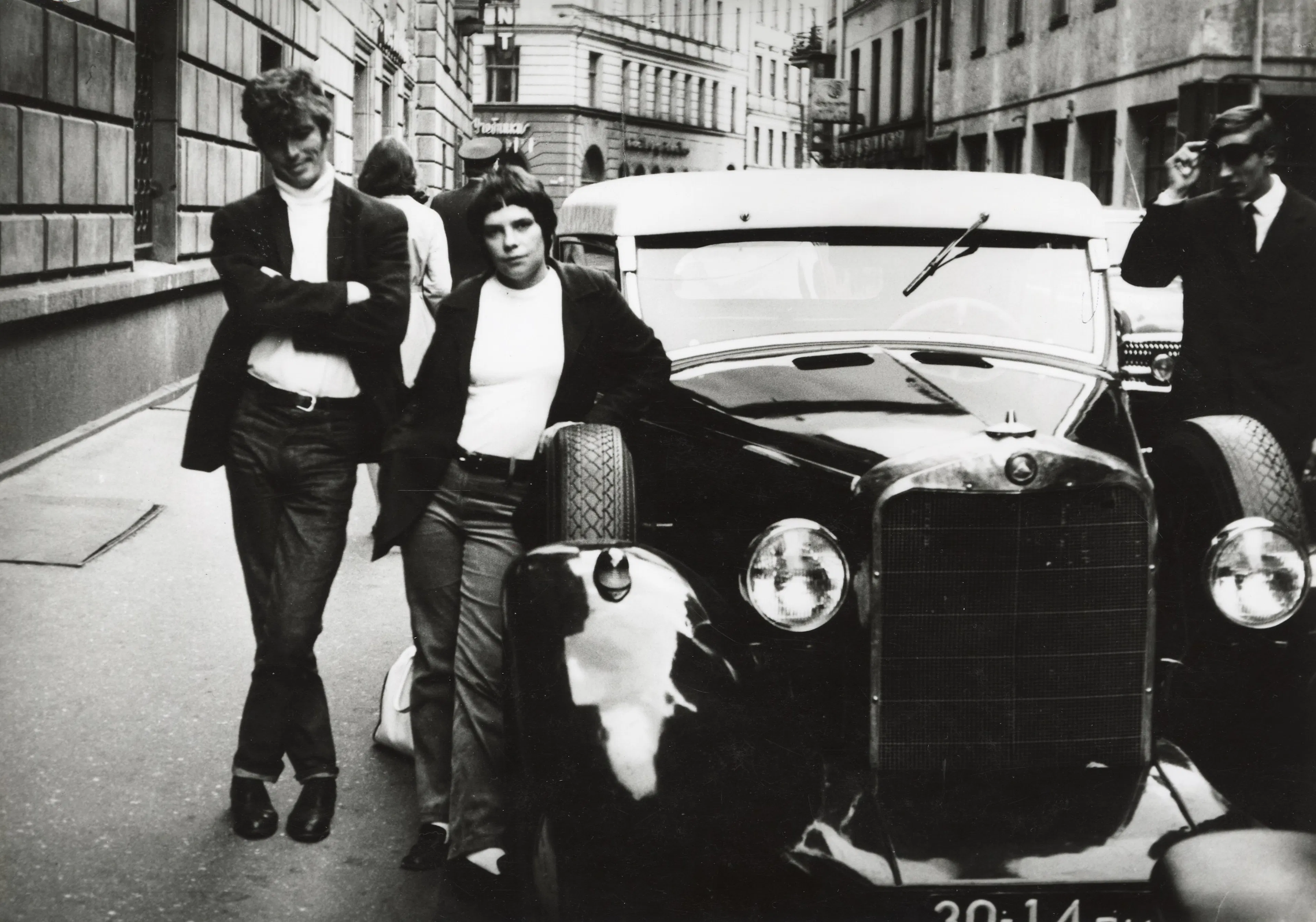 Fotogrāfijā redzams vīrietis un sieviete līdzīgos džemperos un žaketēs. Sieviete ir atspiedusies pret retro automašīnas riepu. Automašīnas kreisajā pusē stāv vīrietis uzvalkā.
