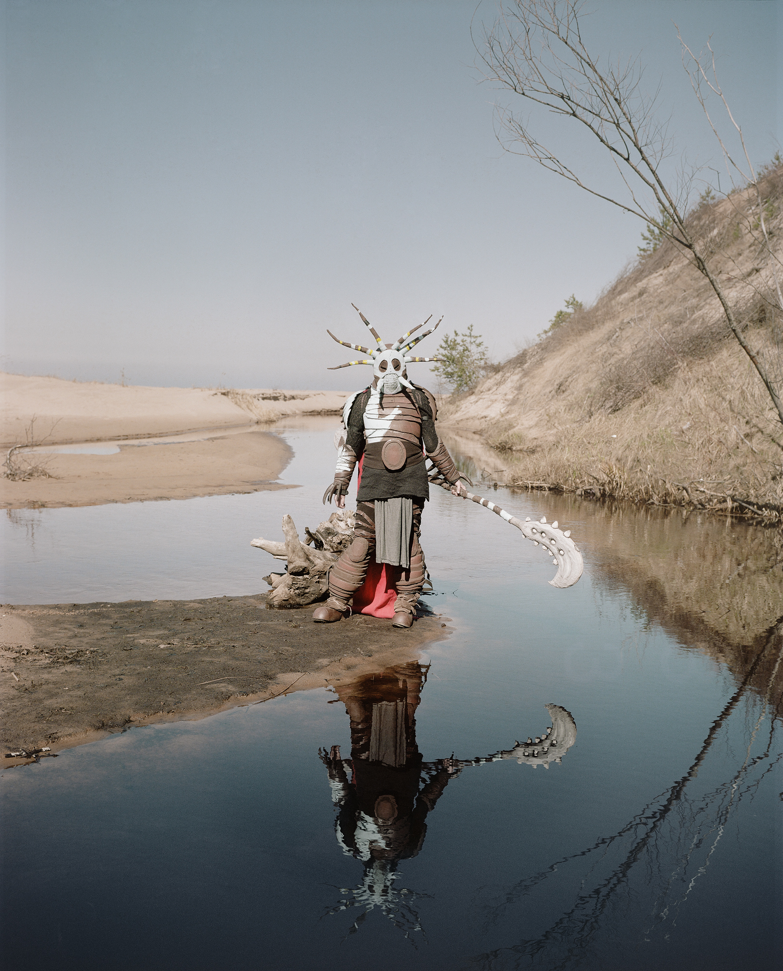 The photograph depicts a person in a fantasy costume with a mask and horn-like headdress, standing in shallow water within a sandy landscape and holding a decorative weapon.
