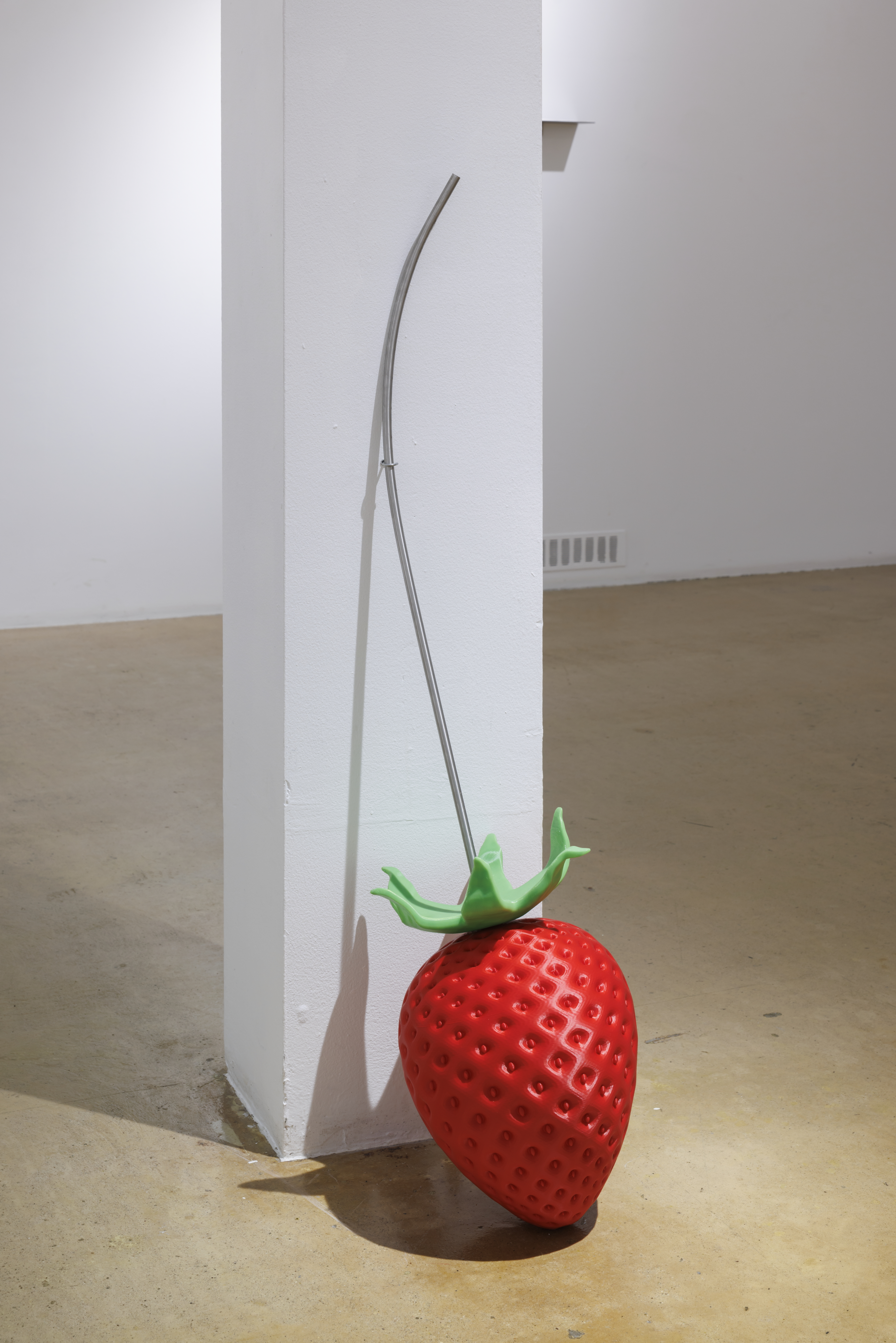 The photograph shows a stylized red strawberry-shaped sculpture with a green leafy crown, from whose top a curved metal rod extends upward; the sculpture is placed beside a white column.
