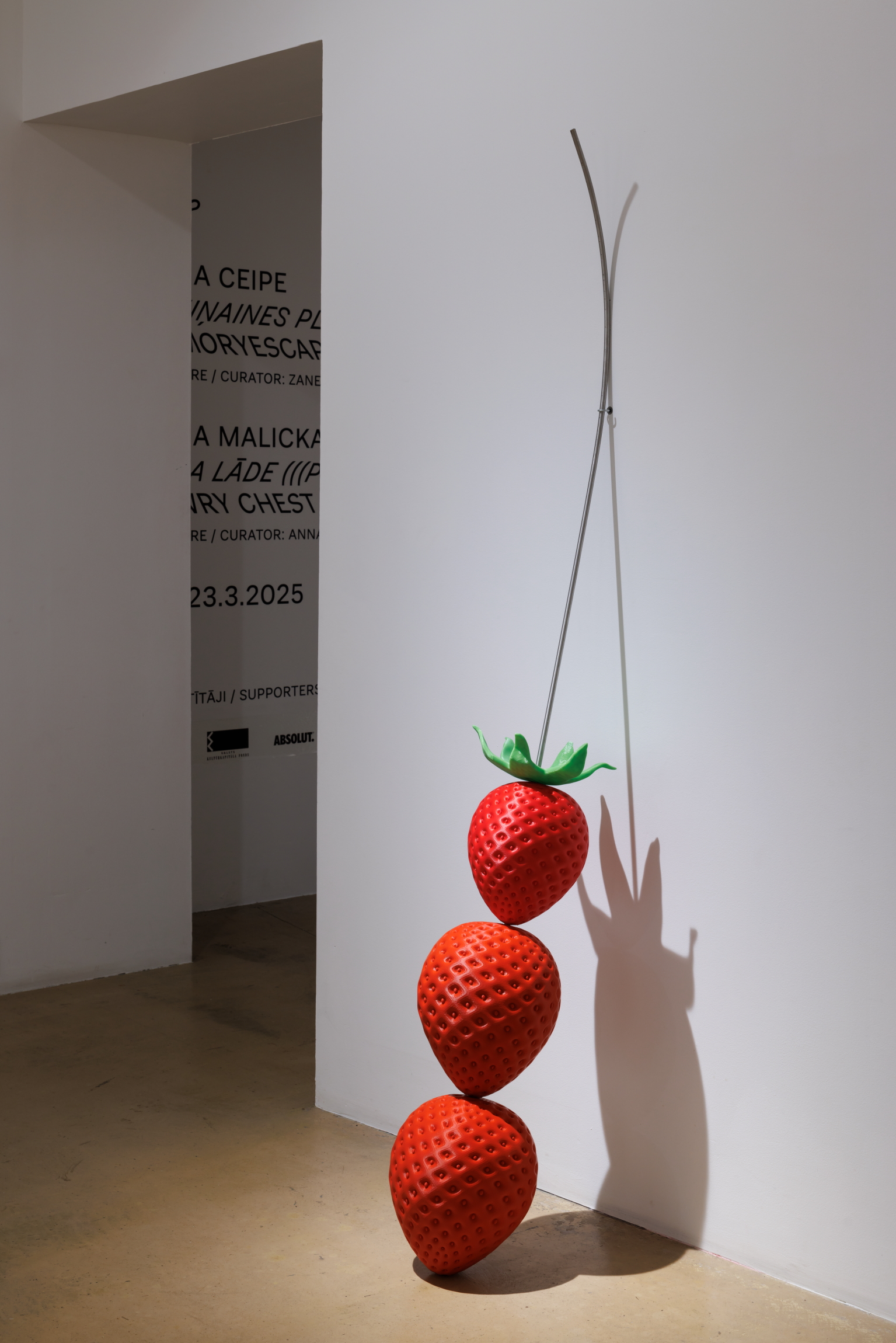 The photograph shows a vertical sculpture composed of three stylized red strawberries stacked one above another and attached to a curved metal rod.
