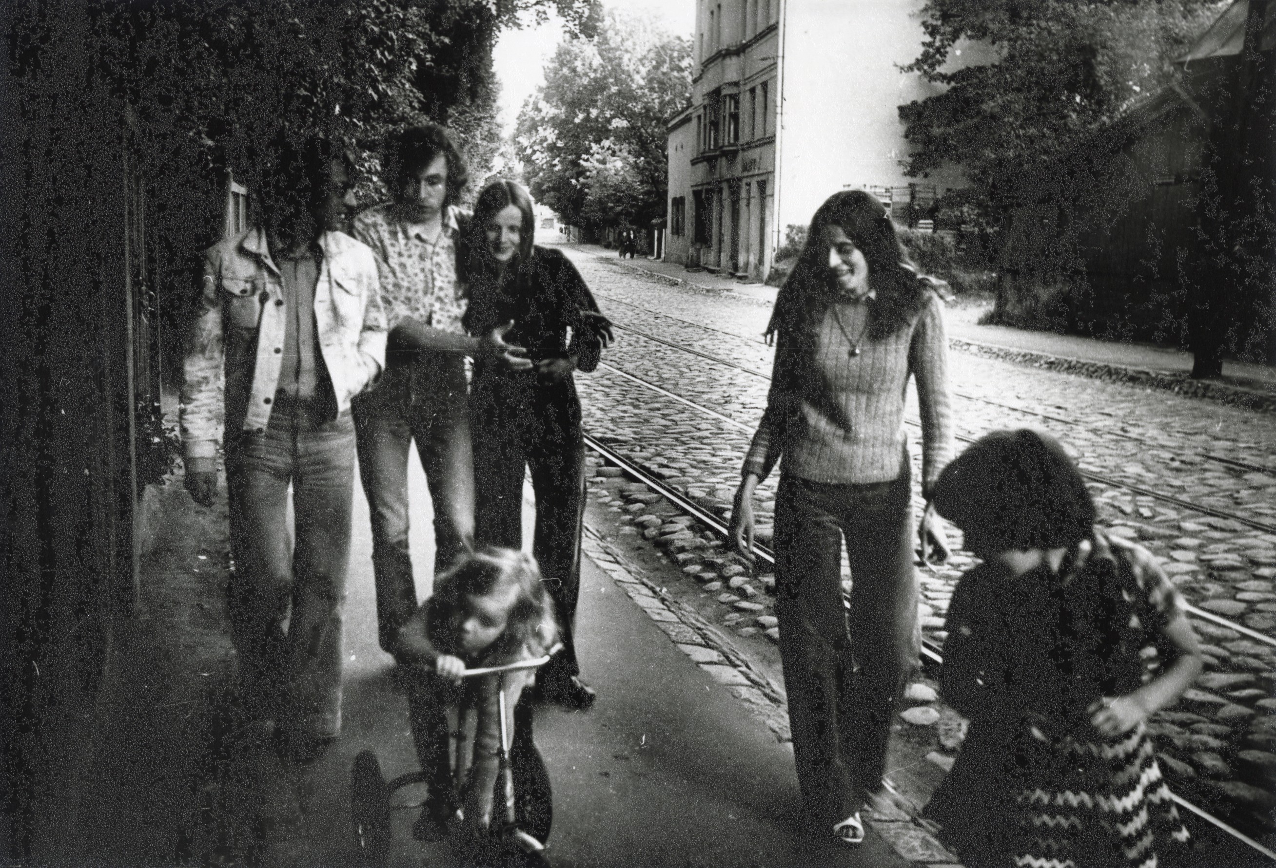Fotogrāfijā redzami jauni cilvēki ar diviem bērniem blakus. Viņi iet pa ietvi F. Brīvzemnieka ielā, blakus redzamas tramvaja sliedes un ēkas.