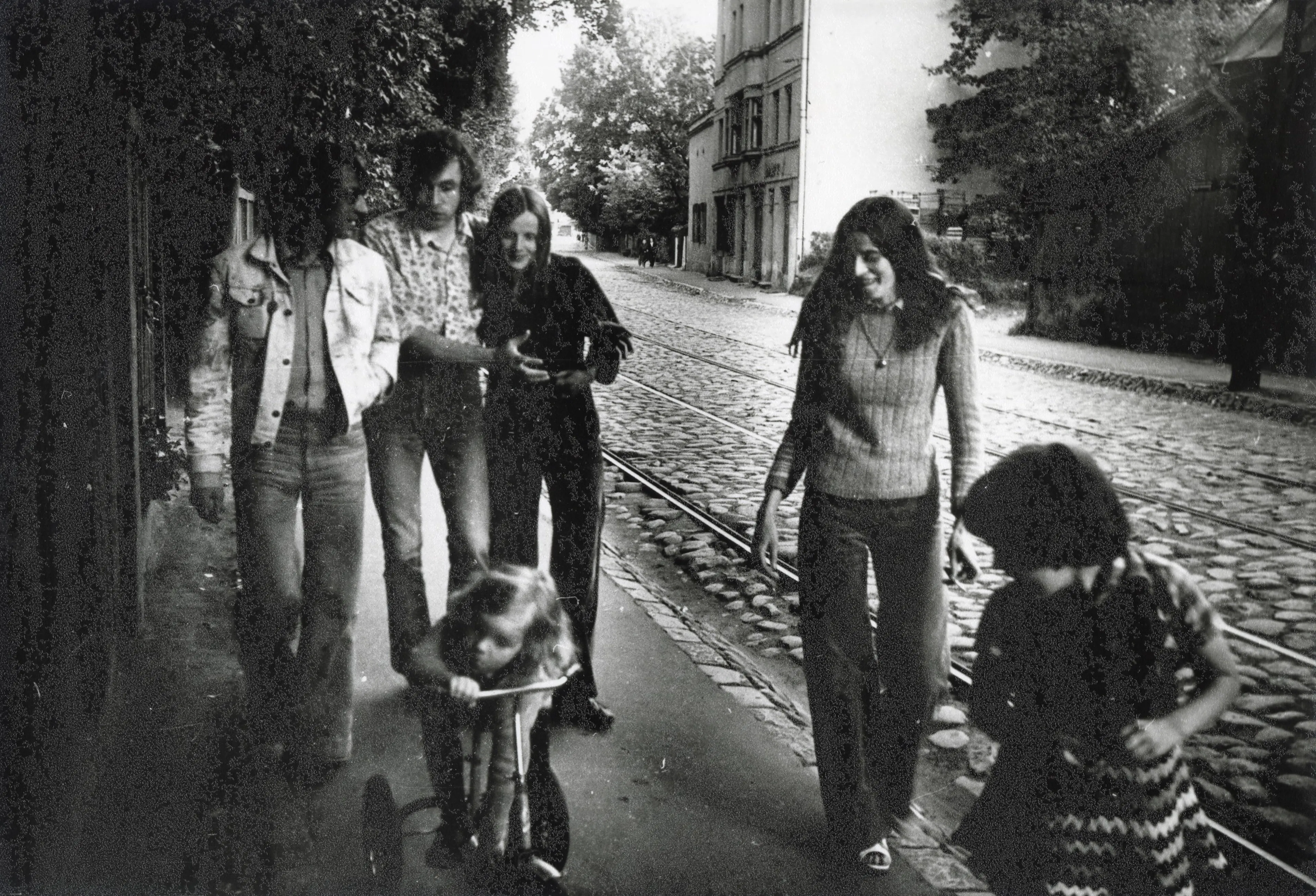 Fotogrāfijā redzami jauni cilvēki ar diviem bērniem blakus. Viņi iet pa ietvi F. Brīvzemnieka ielā, blakus redzamas tramvaja sliedes un ēkas.