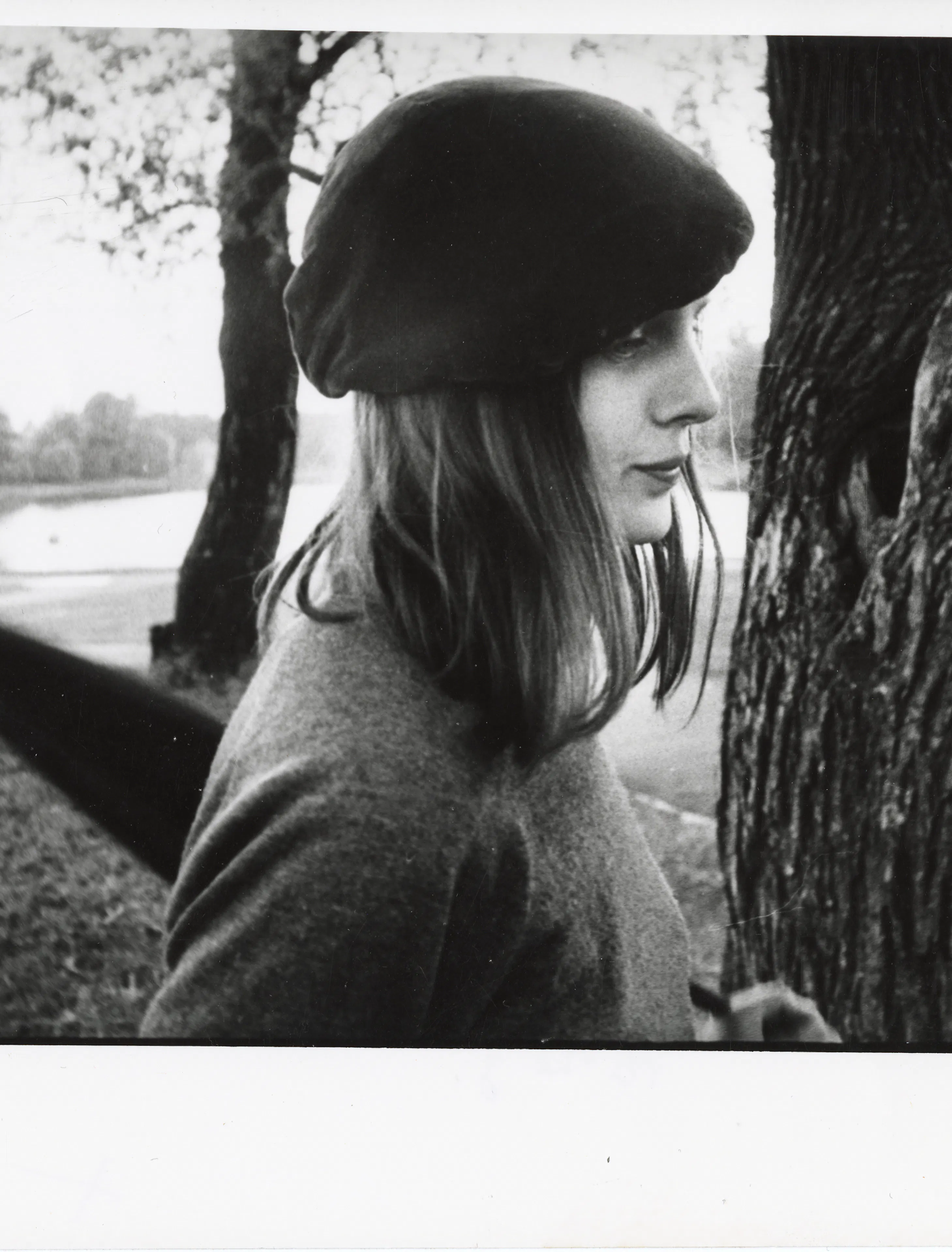 The photograph shows a woman in a beret standing near a tree trunk. In the background is Māra's pond.