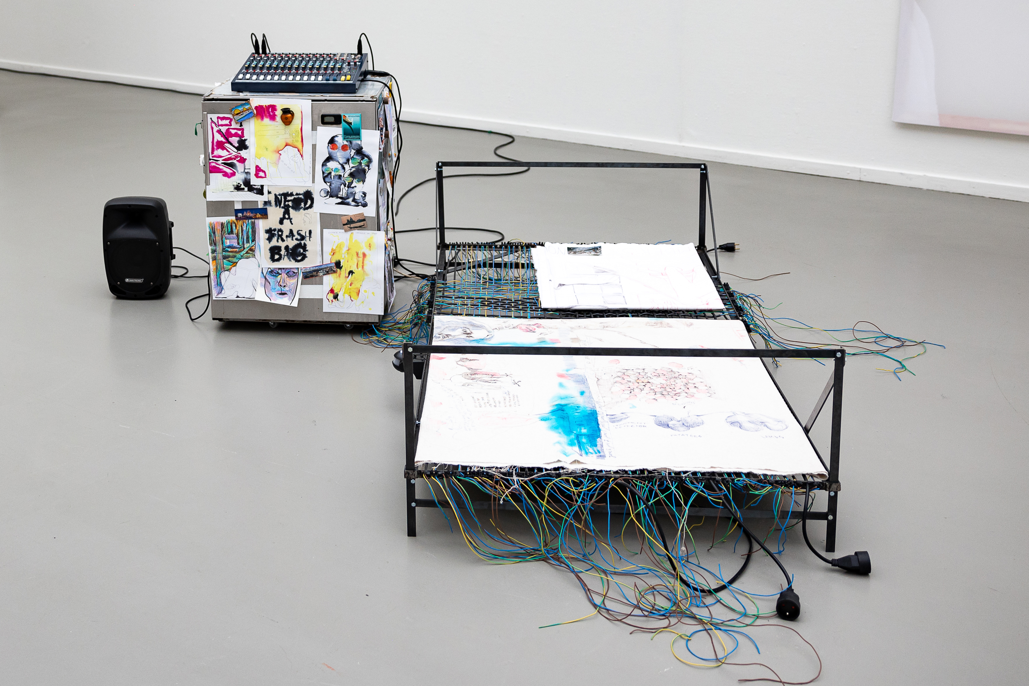 At the centre is a metal bed frame with fabrics covered in drawings and numerous colourful wires. Beside it is a box covered with drawings and images, topped with an audio mixer, as well as a black speaker.