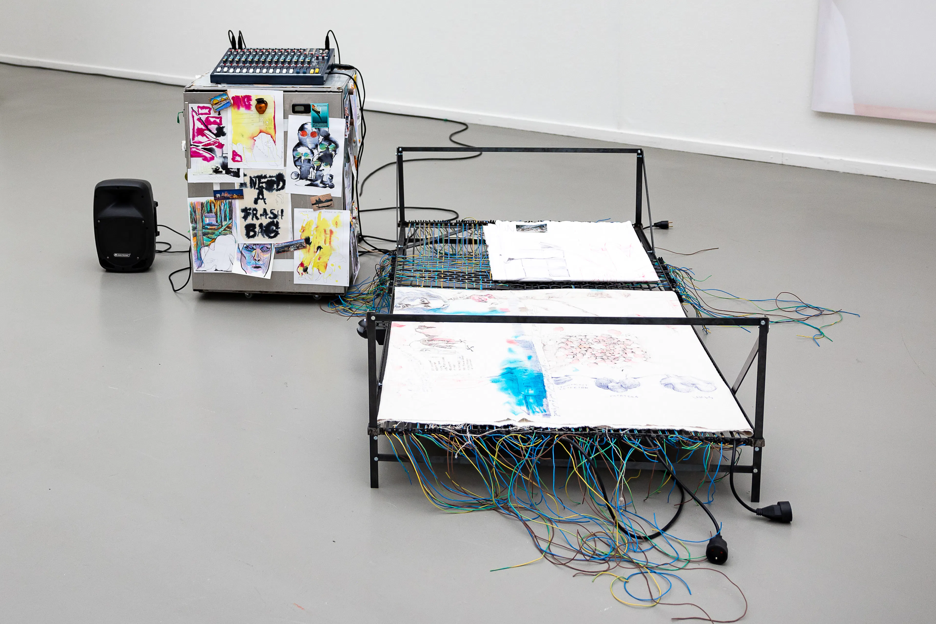 At the centre is a metal bed frame with fabrics covered in drawings and numerous colourful wires. Beside it is a box covered with drawings and images, topped with an audio mixer, as well as a black speaker.