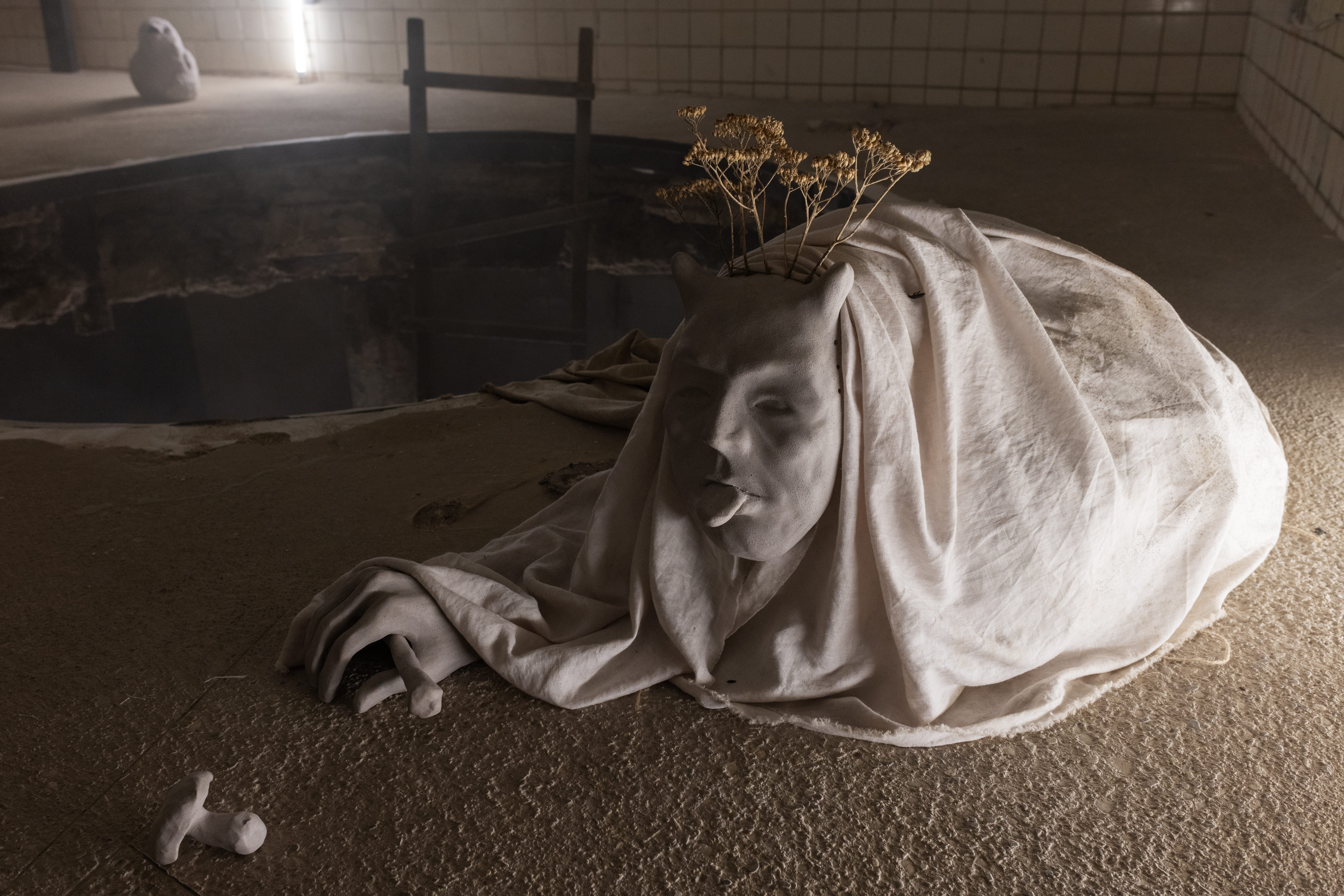 A sculptural, figurative object consisting of several parts. A ceramic mask is attached with a cord to sandbags covered with textile, creating the illusion of a crawling body holding a bone in one hand and reaching towards a nearby ceramic mushroom.