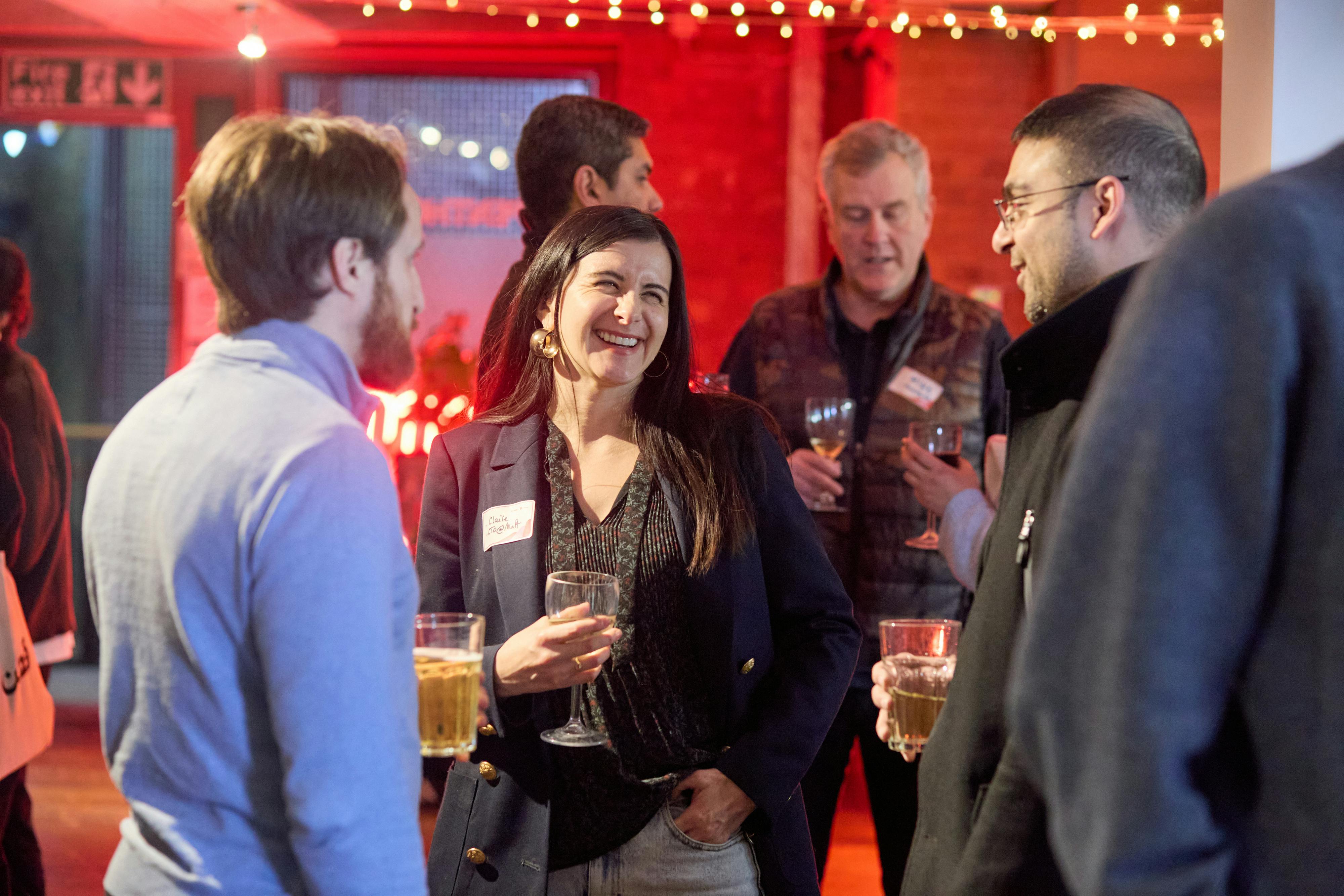 A lively networking event with professionals chatting over drinks. A woman in a dark blazer laughs while engaging in conversation.