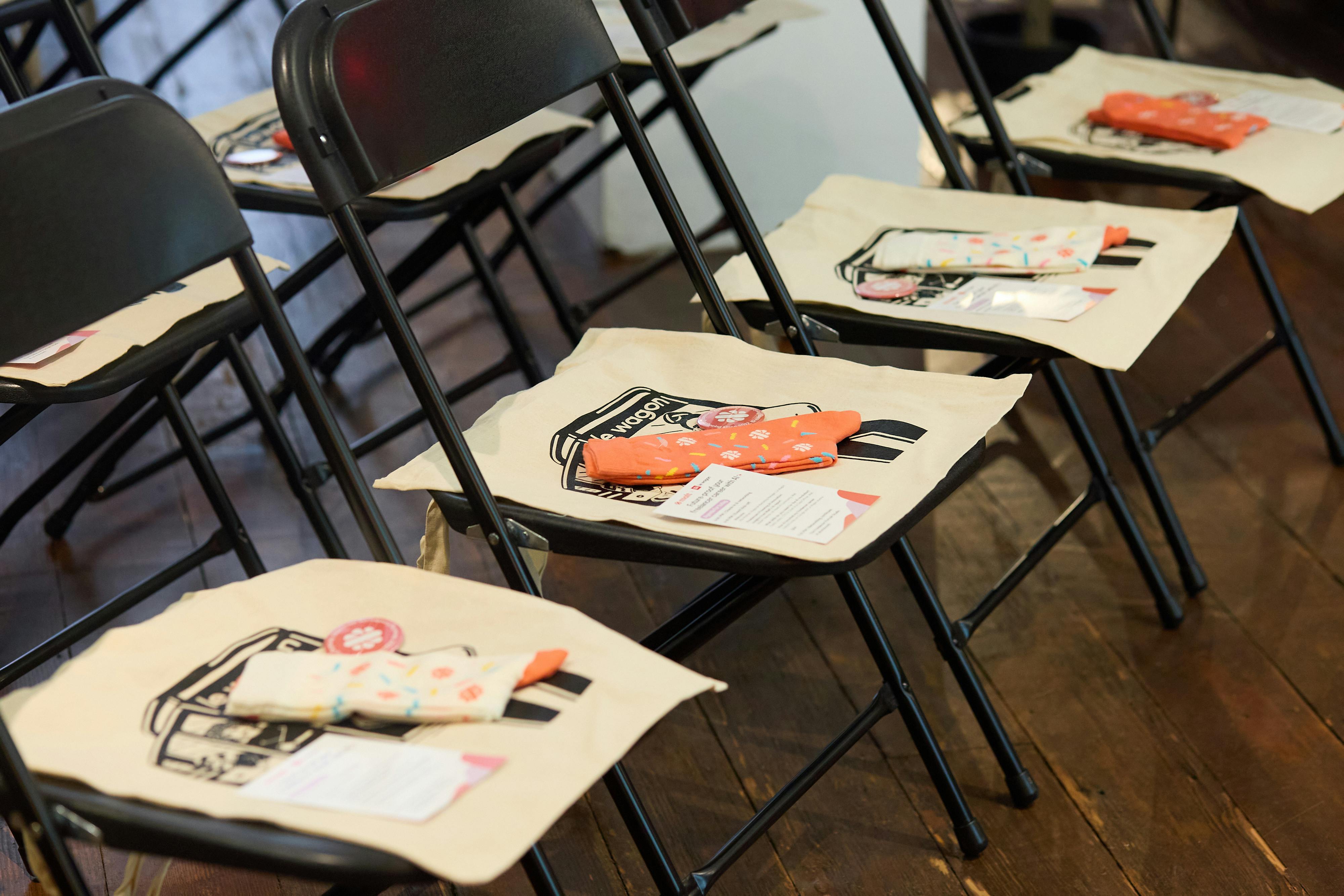 A row of black folding chairs with tote bags, socks, and event materials neatly placed on each seat, ready for attendees.