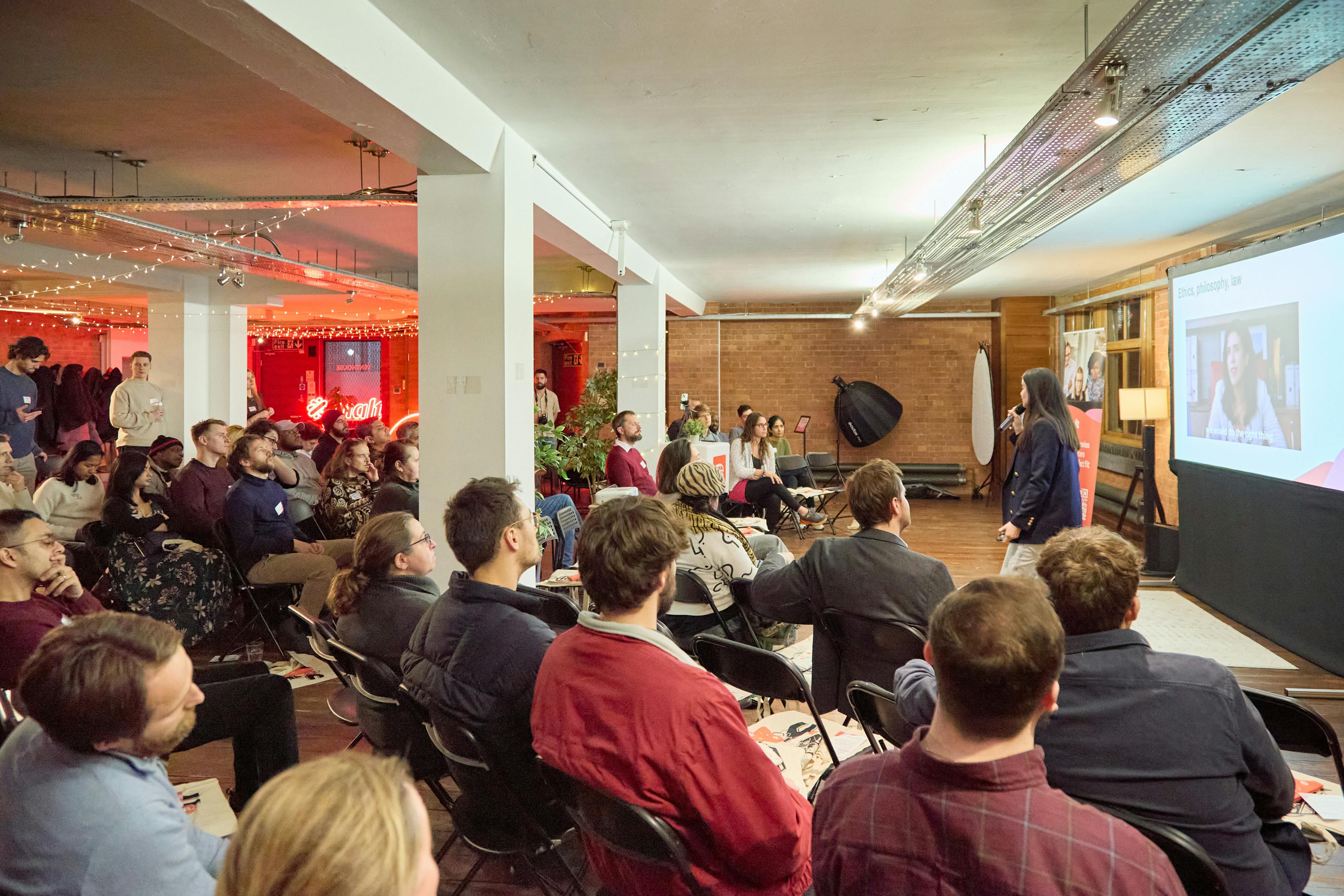  A packed event space with attendees seated and engaged in a presentation. A speaker stands at the front, addressing the audience with a projected slide behind them. The room is warmly lit with string lights.