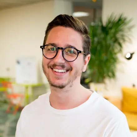 Portret van een jonge blank man met een wit shirt en een bril die lachend in de camera kijkt met op de achtergrond een kantooromgeving 