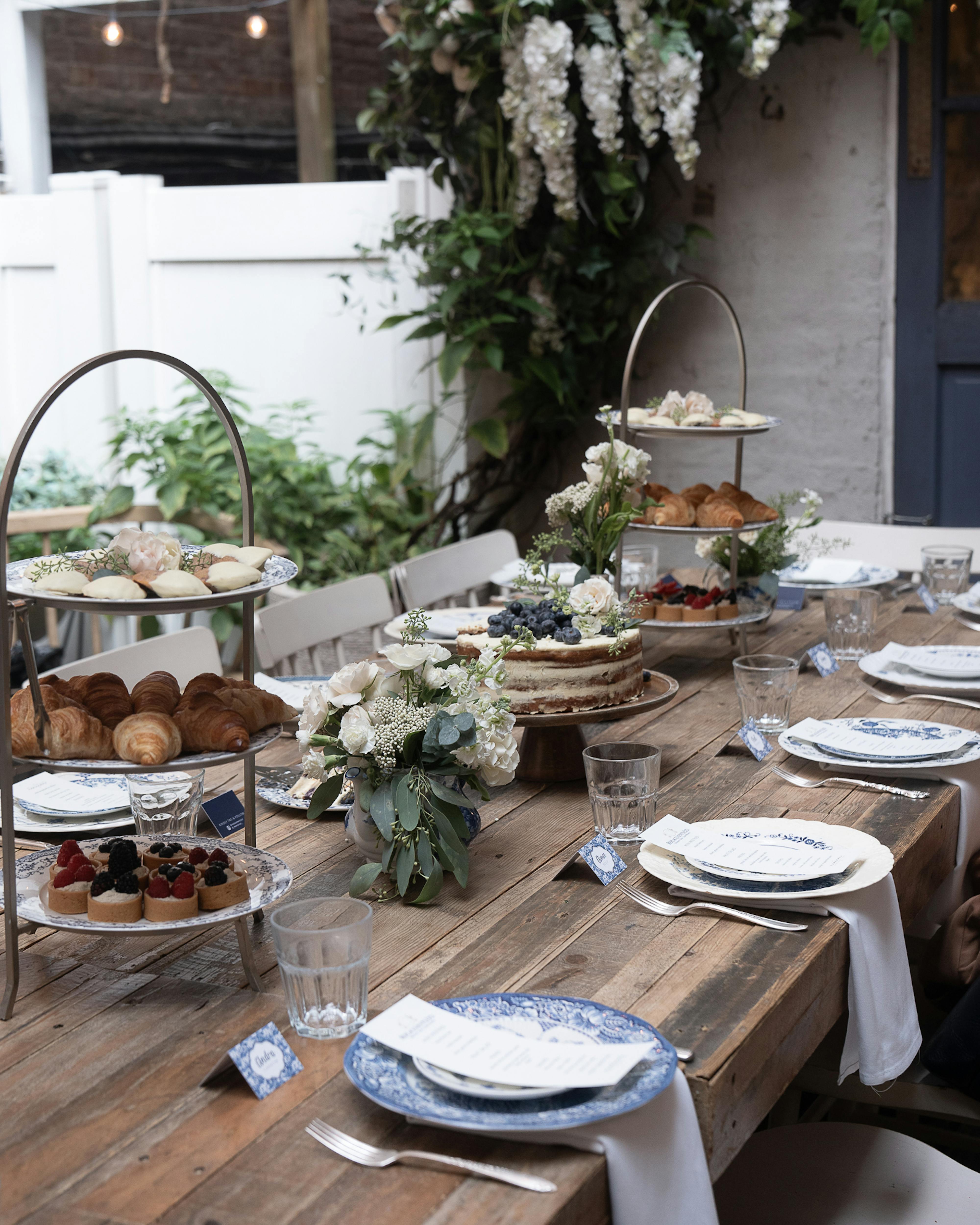 long rustic wooden table with blue and white plates and table settings, two tiered stands with pastries, and a rustic vanilla cake in the middle of the table