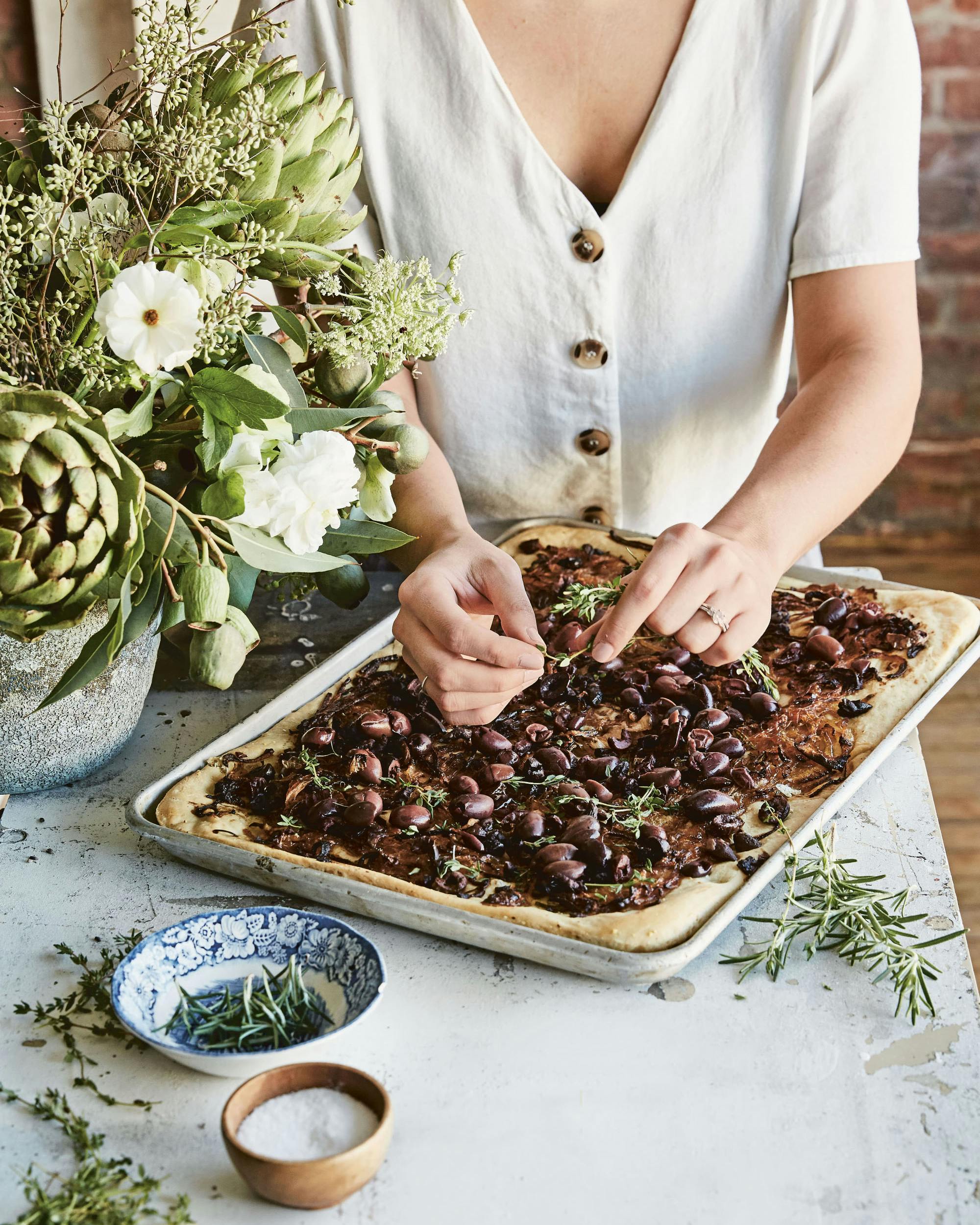 Hands placing herbs on Pissaladiere from maman the cookbook