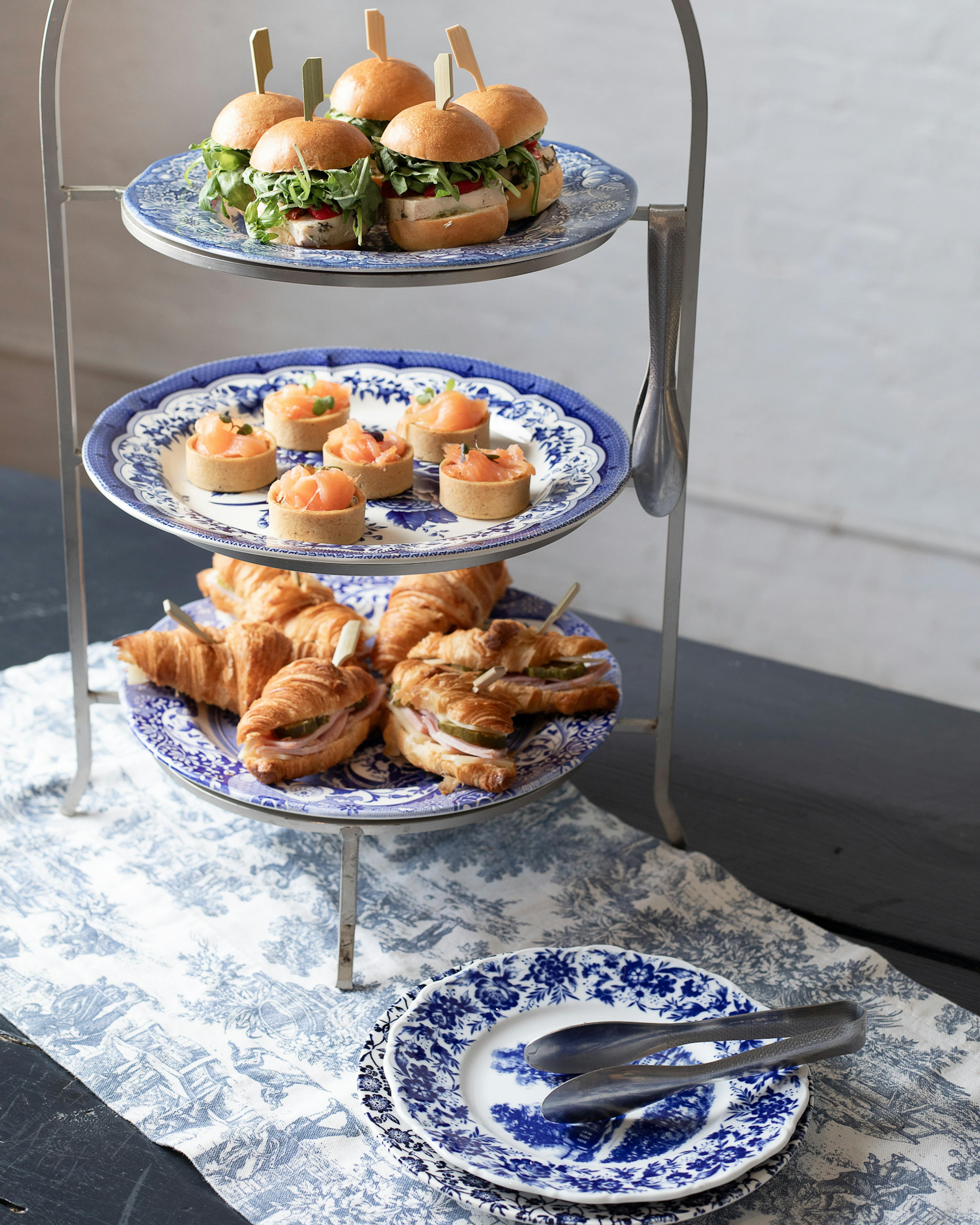 a tiered food stand with mini sandwiches, tarts, and blue and white plates