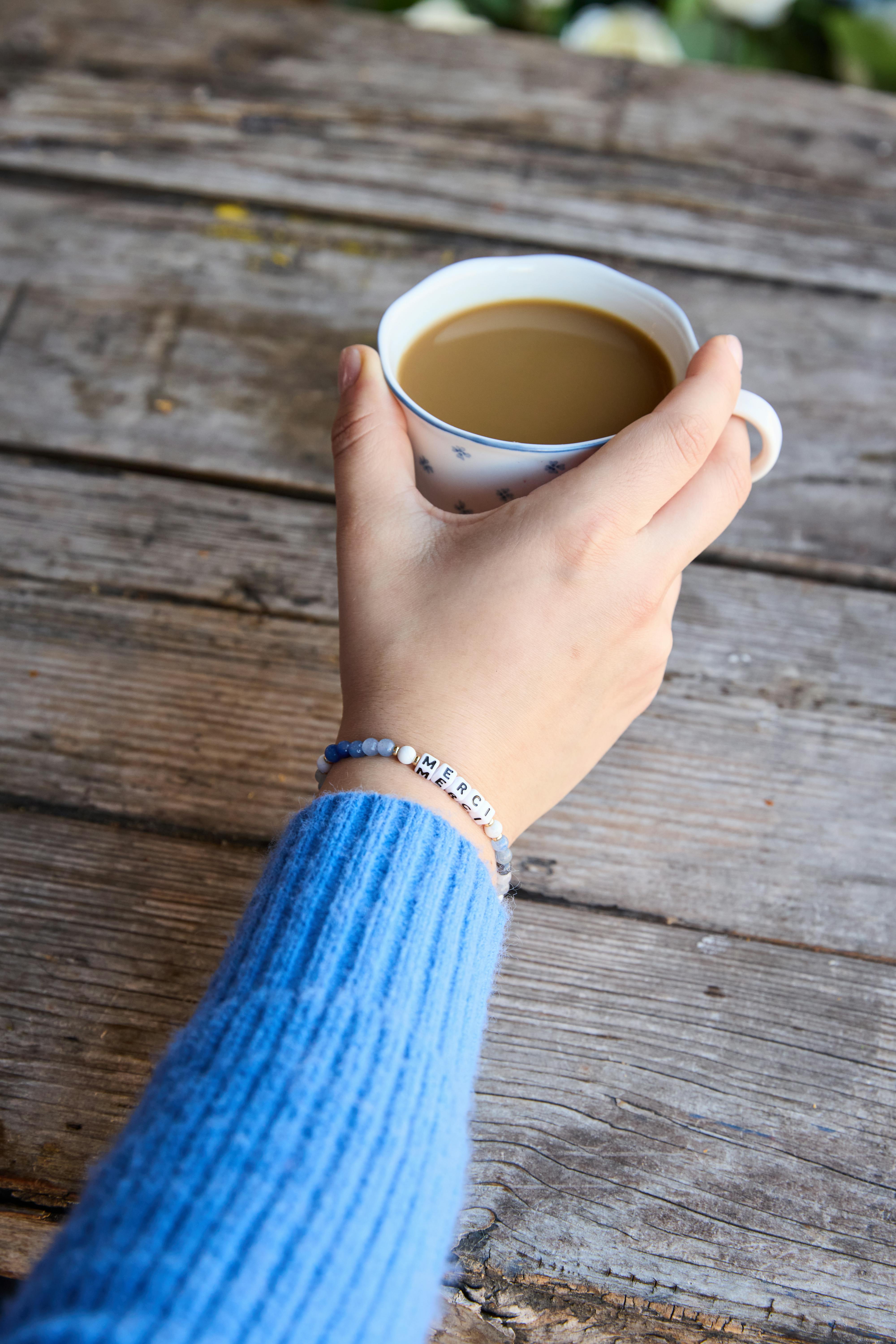 close-up of a hand wearing a blue beaded bracelet that spells "merci," holding a cup of coffee in a white mug with small blue flowers, resting on a rustic wooden table